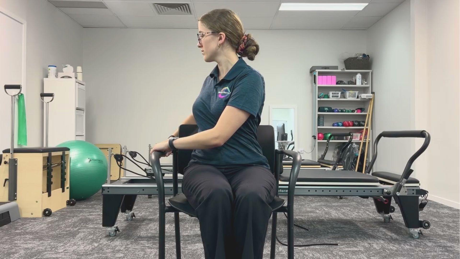 Woman in a physio polo sits on a chair, rotating her torso to one side in a clinic setting.