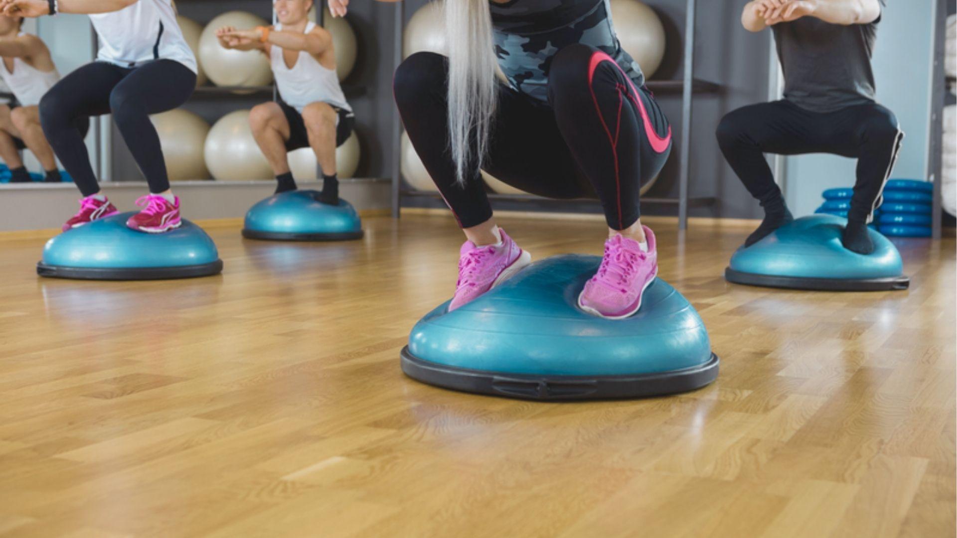 Group fitness class performing squats on blue BOSU balls in a gym with mirrors and exercise balls.