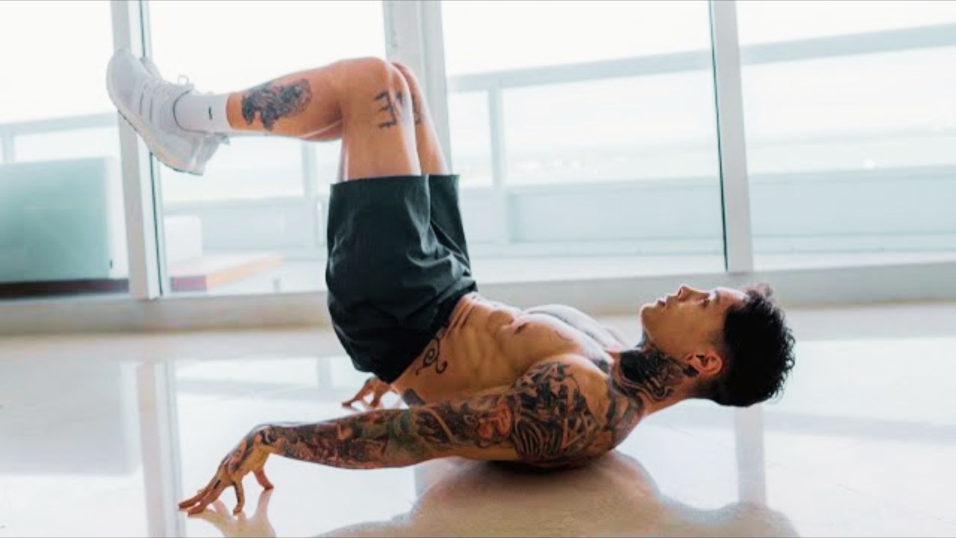 A tattooed man lies on his back on a bright floor with legs raised and knees bent, balancing on his upper back.