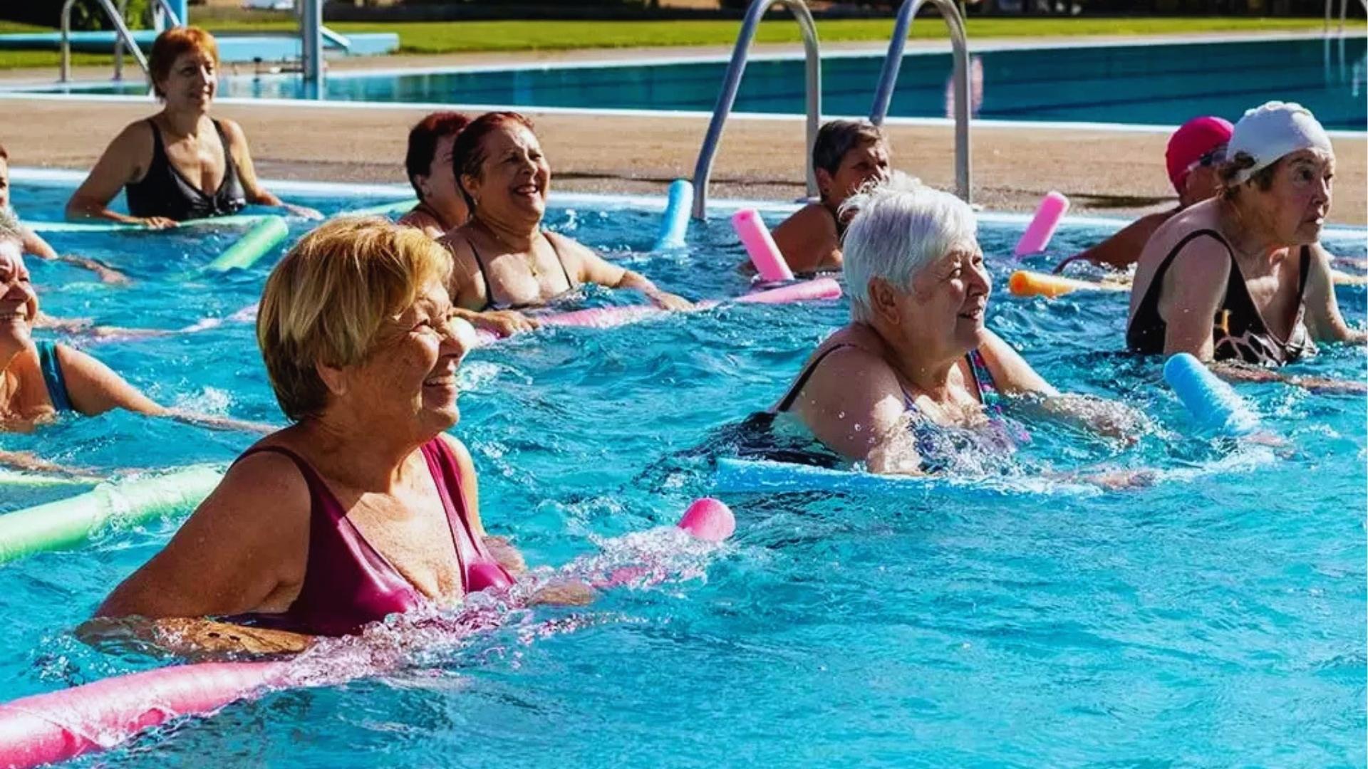 Senior women smile during water aerobics class using colorful pool noodles in an outdoor pool for joint-friendly exercise.