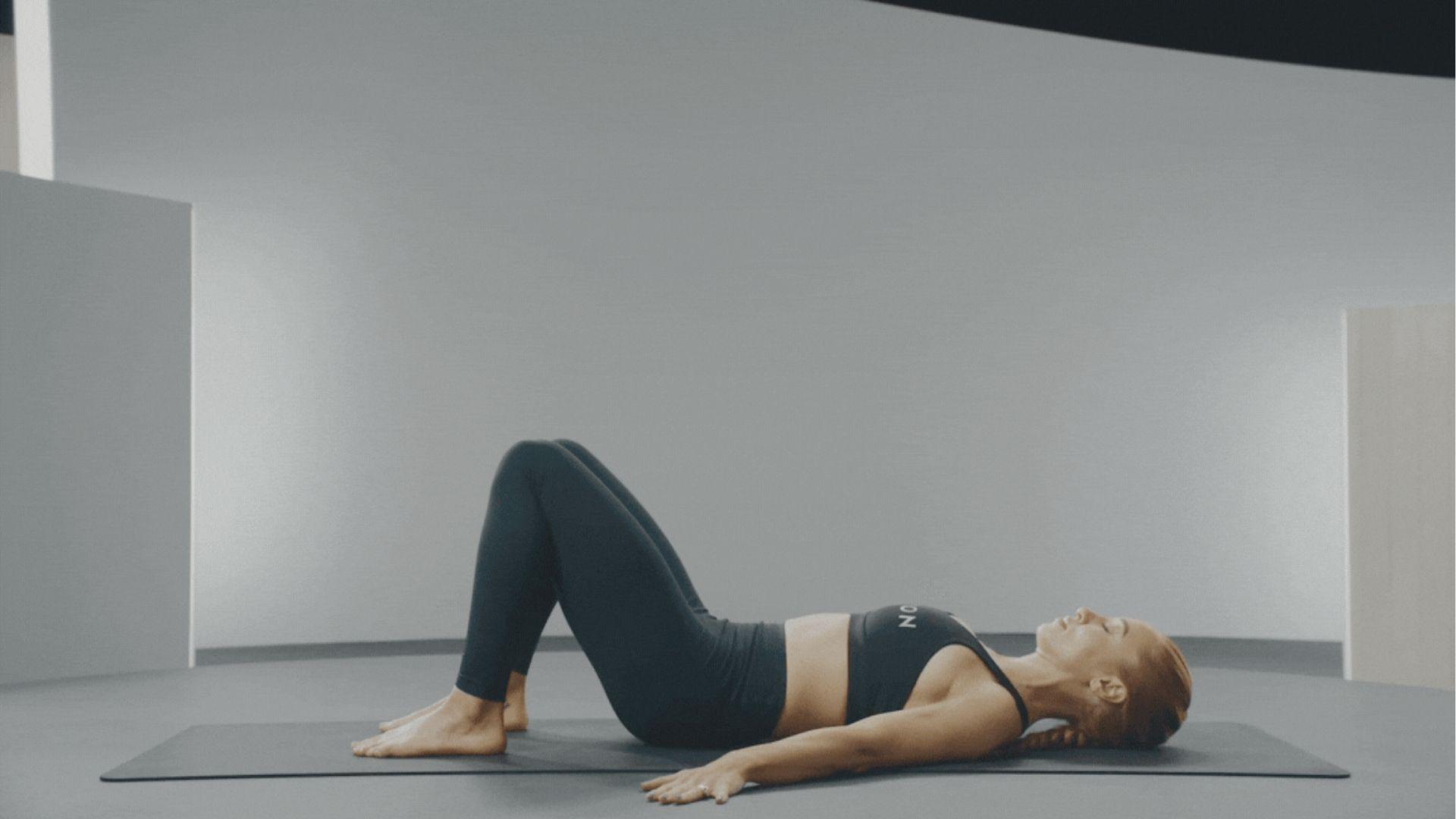 Woman in black activewear lying on a gray yoga mat with knees bent, preparing for a pelvic tilts.
