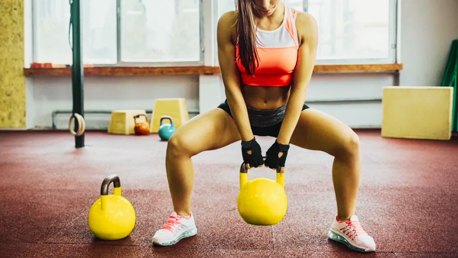 Woman in a fitness studio performing a sumo squat with a yellow kettlebell held between her legs.