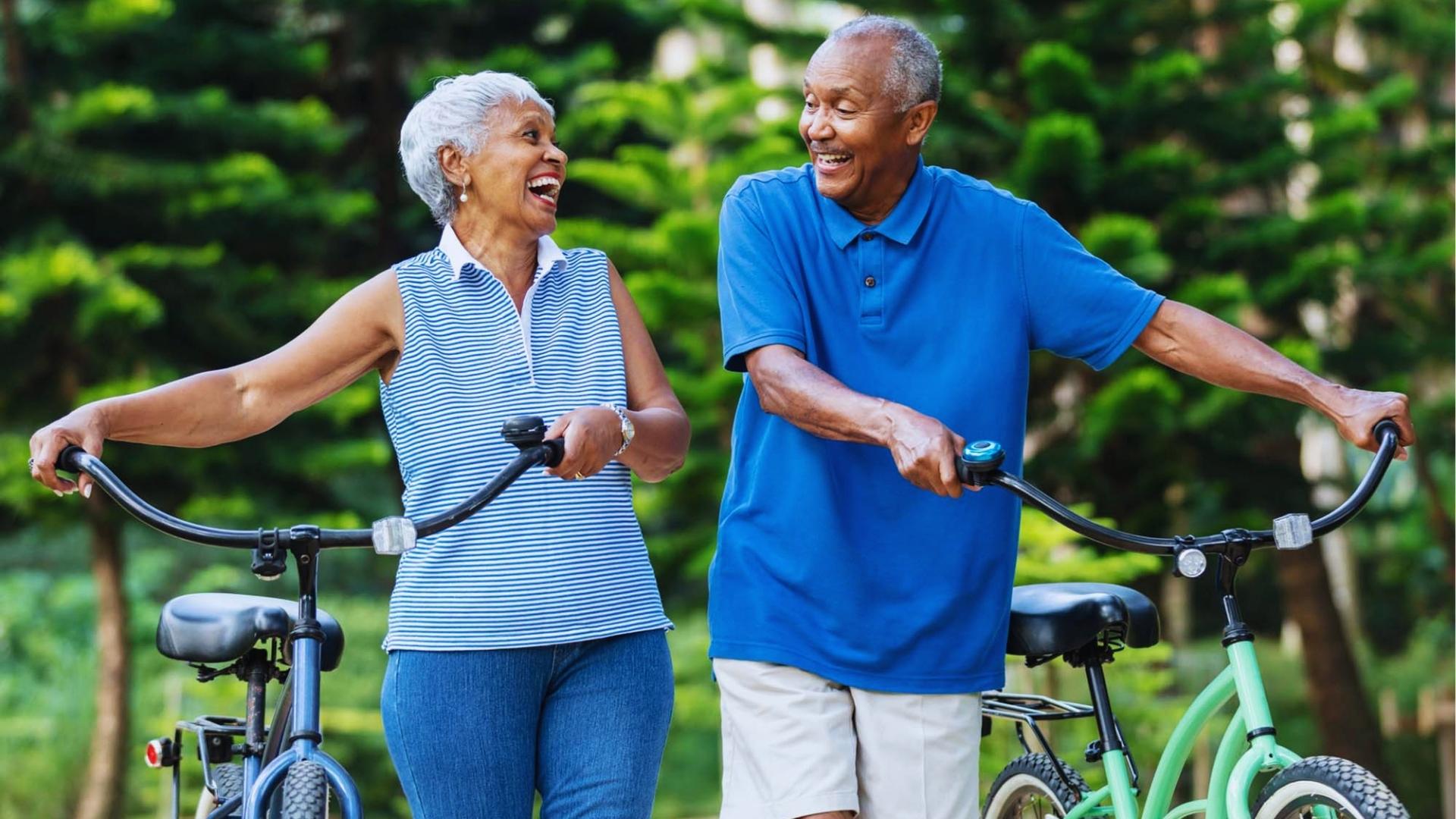 Happy senior couple walks side by side with cruiser bikes on sunny park path for low-impact cardio.