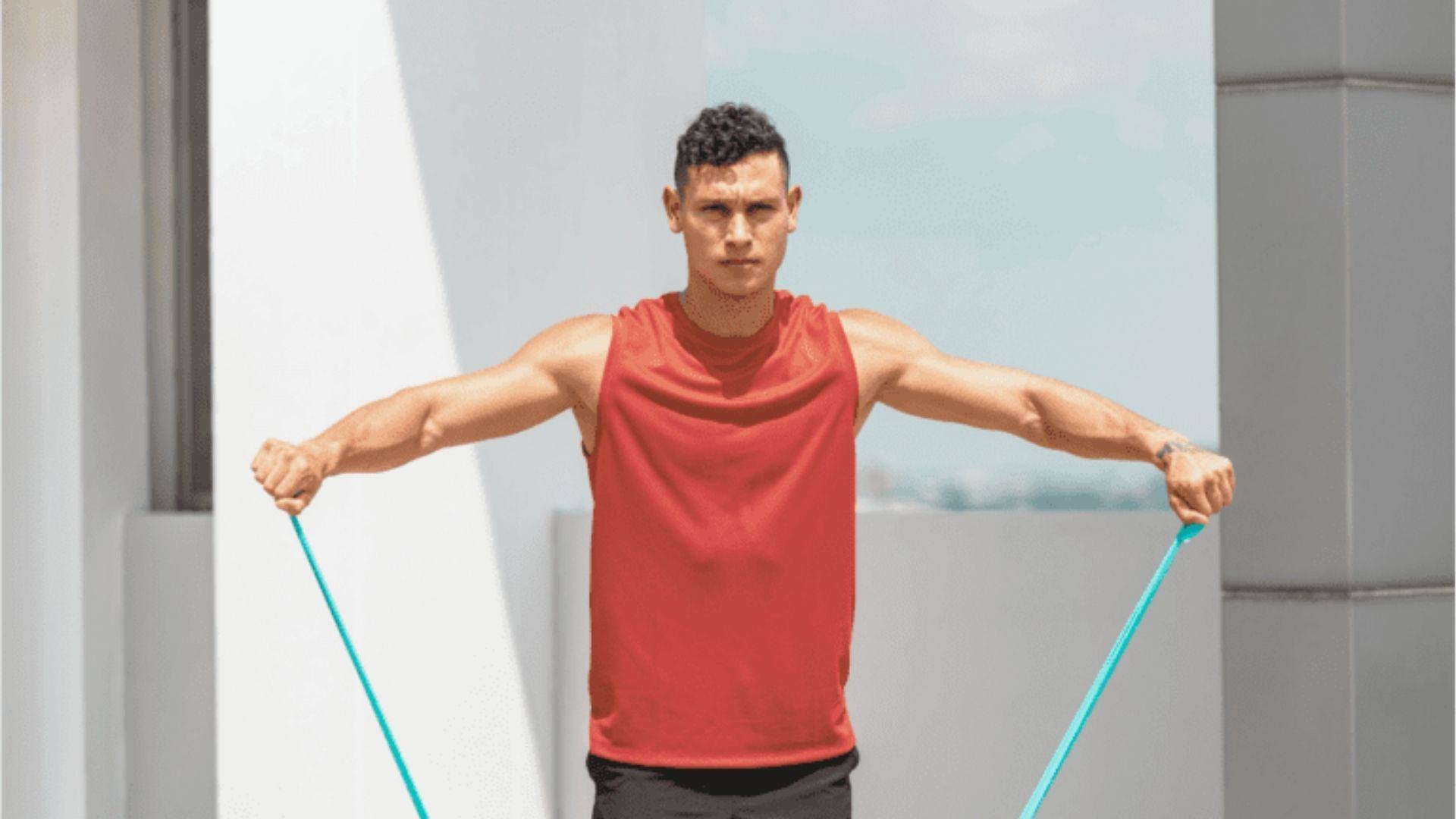 A man in a red sleeveless shirt performs a resistance band lateral raise outdoors.