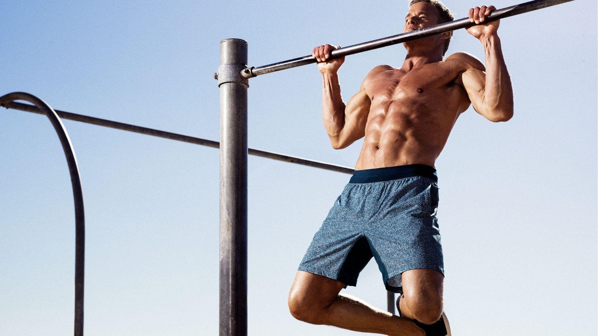 Shirtless man with athletic build executing an outdoor pull-up mid-rep