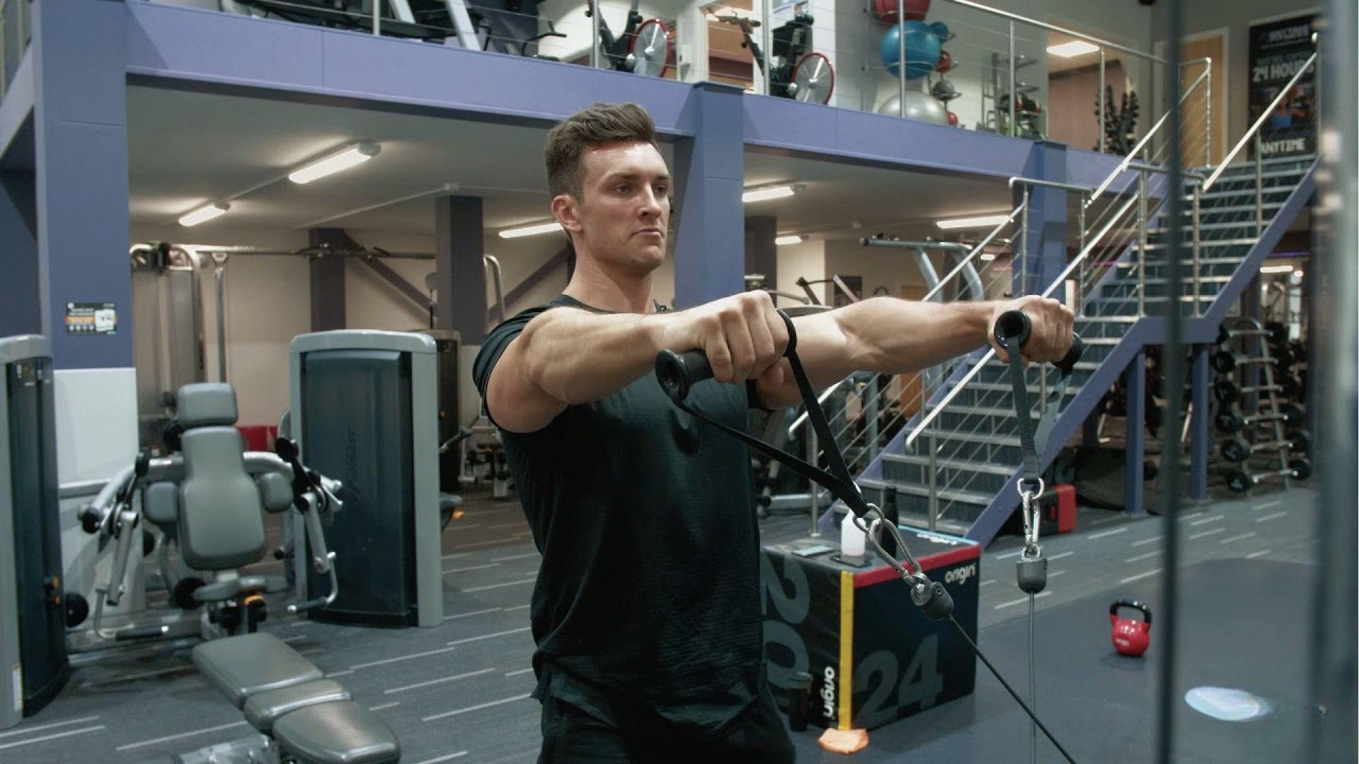A man in black shirt executes cable Y raise holding handles with arms angled up in the gym.