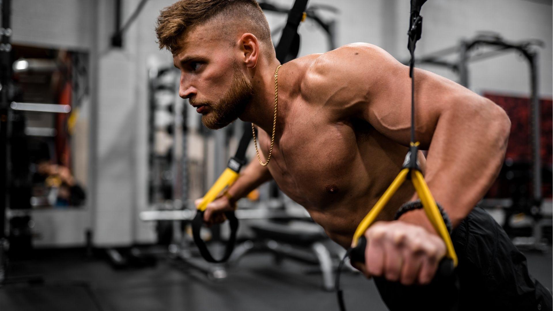 Shirtless man with a gold chain performs a challenging TRX push-up using suspension straps.