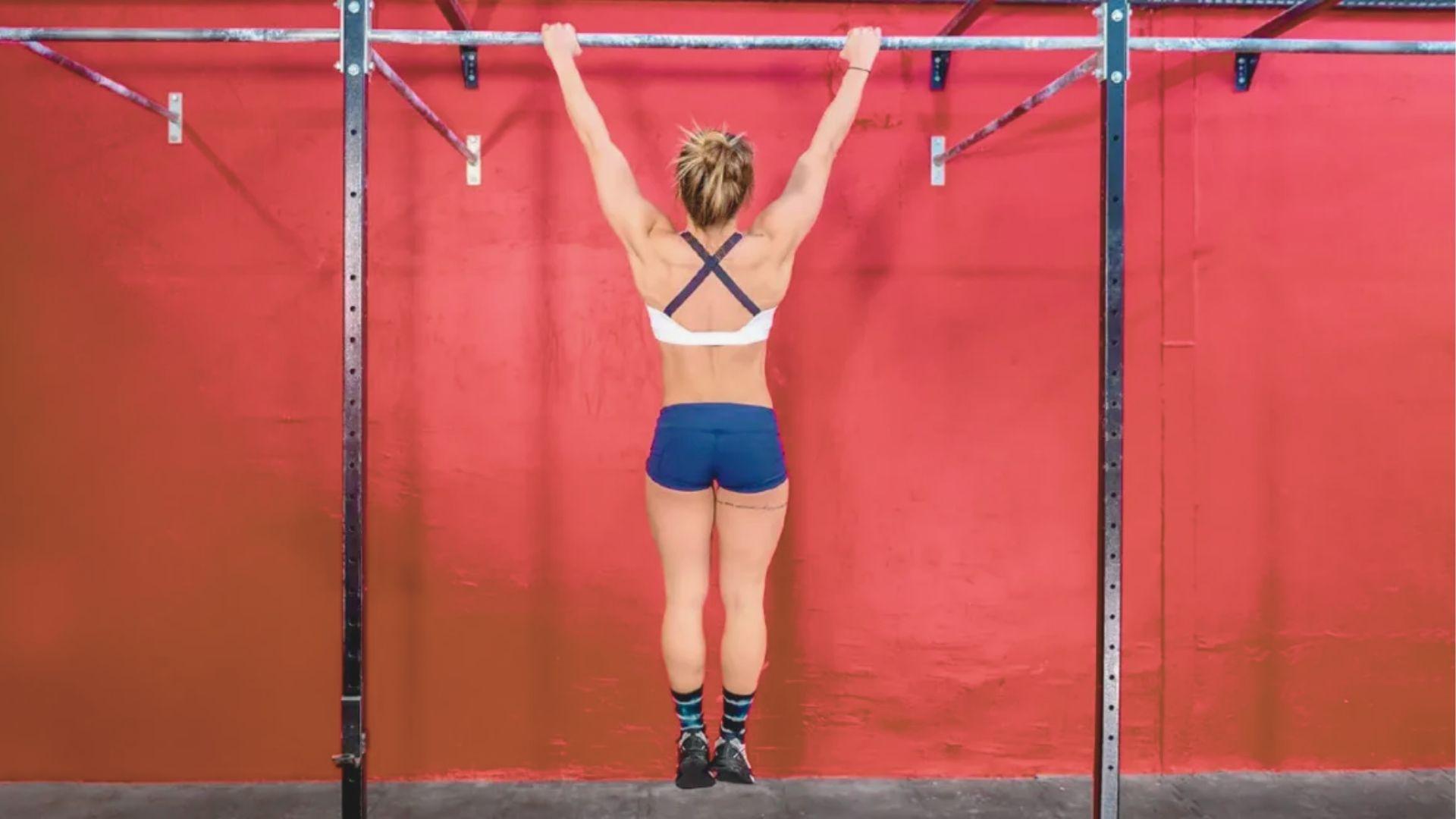 Woman in white sports bra and blue shorts doing a dead-hang pull-up facing a red wall.