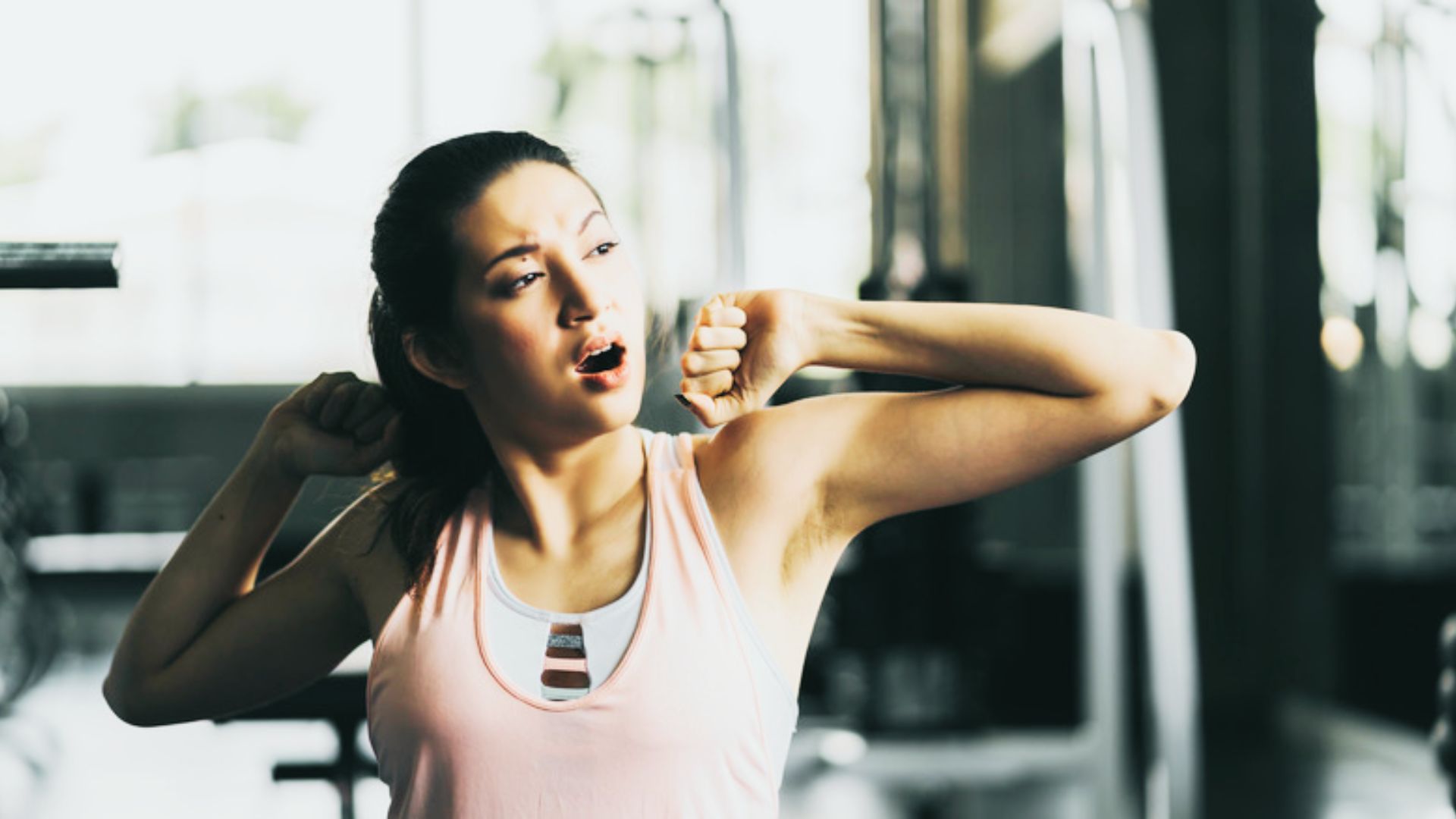 A woman in a pink tank top stretches her arms while yawning, looking sleepy in a gym setting.