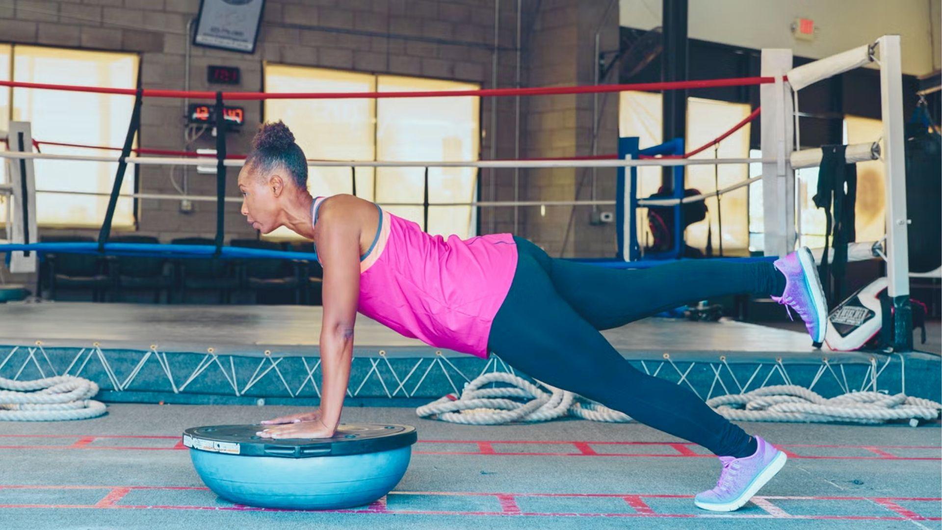 Woman in pink top holds a single-leg plank with hands on a BOSU ball near a boxing ring.