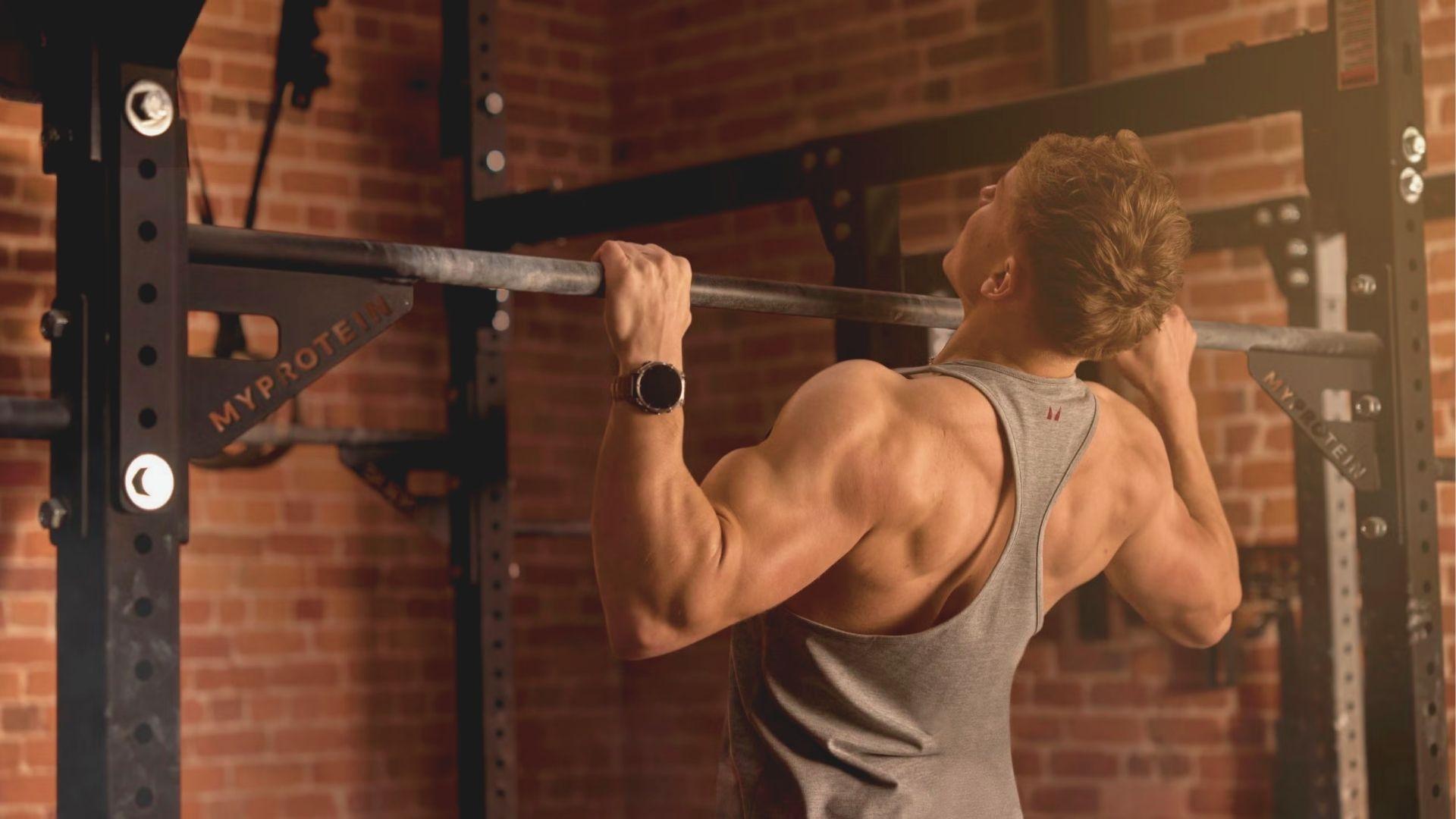 Muscular man in gray tank top performing a pull-up on a MYPROTEIN rack.