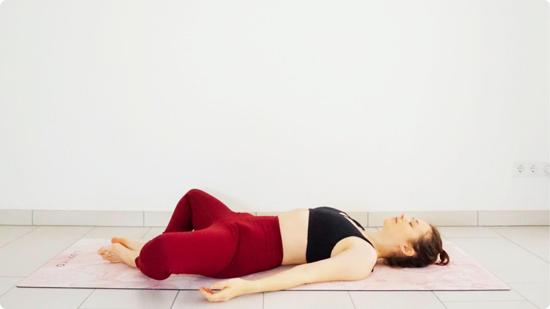 Woman in maroon leggings and black sports bra lying on a pink mat in Reclining Bound Angle Pose.