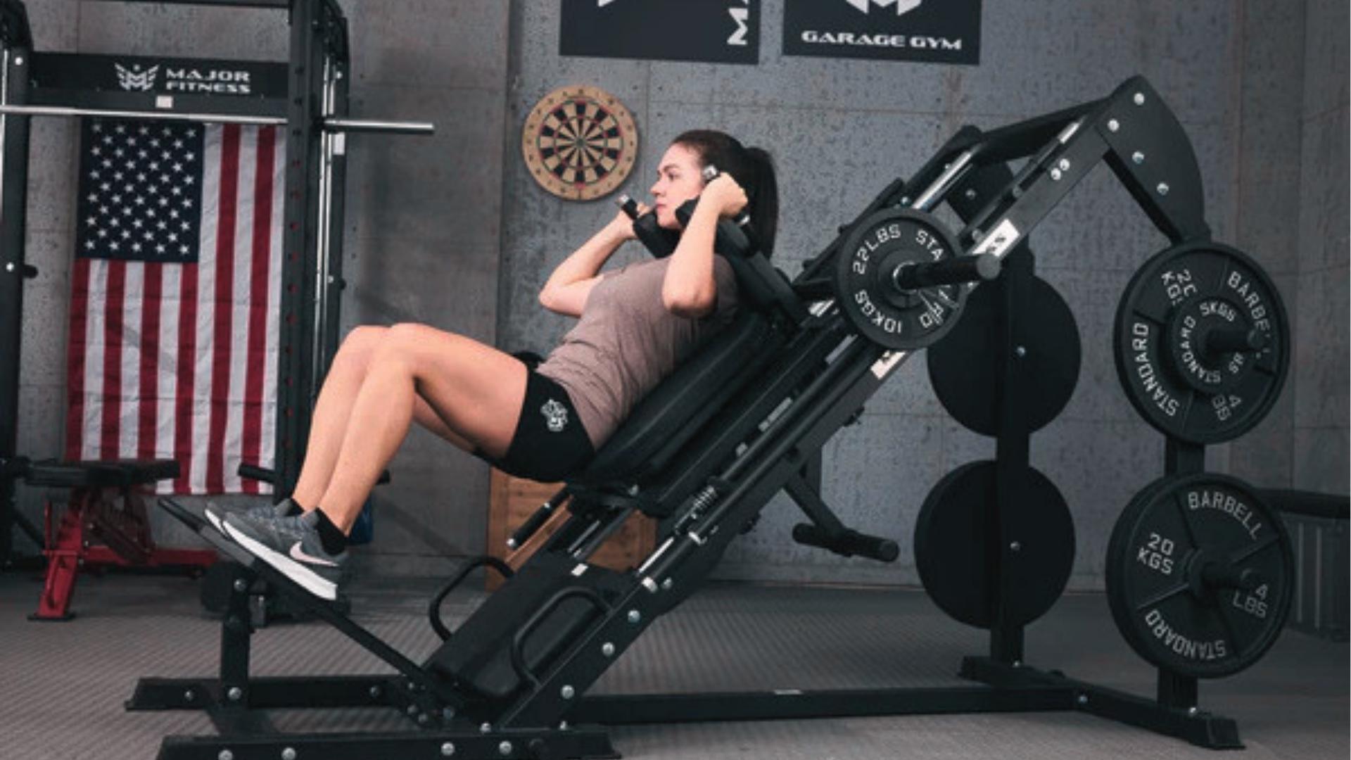 Woman in brown tee and black shorts uses angled hack squat machine in garage gym with American flag backdrop.
