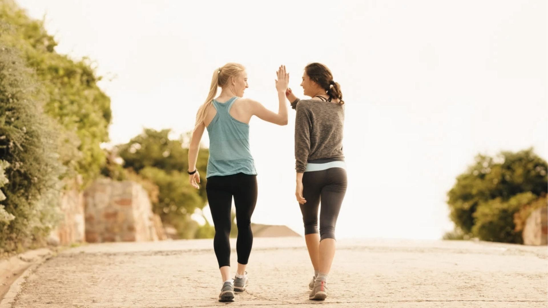 Two women in leggings and tanks high-five while walking on a sunny path, celebrating fitness and step goals.