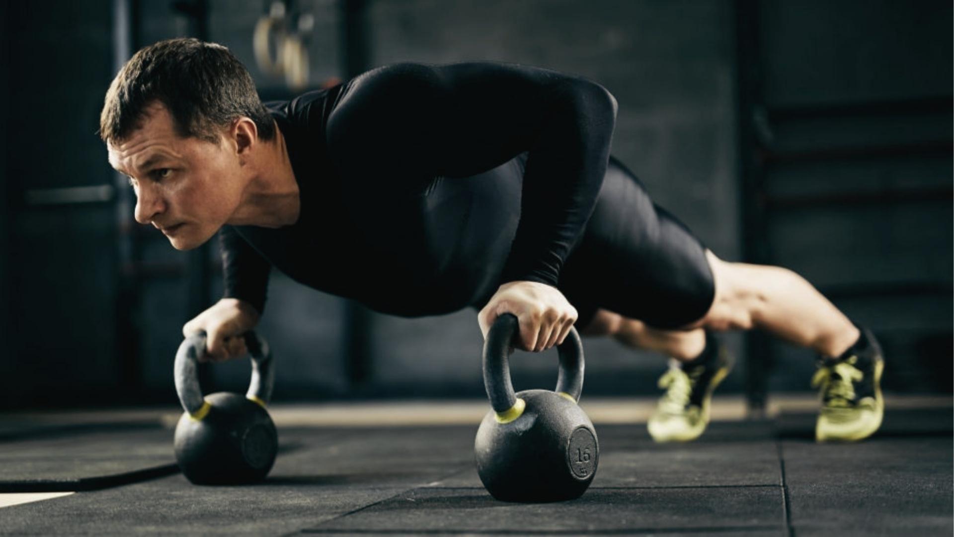 Focused man in black long-sleeve shirt does deficit push-ups on kettlebells in dark gym, increasing chest range.