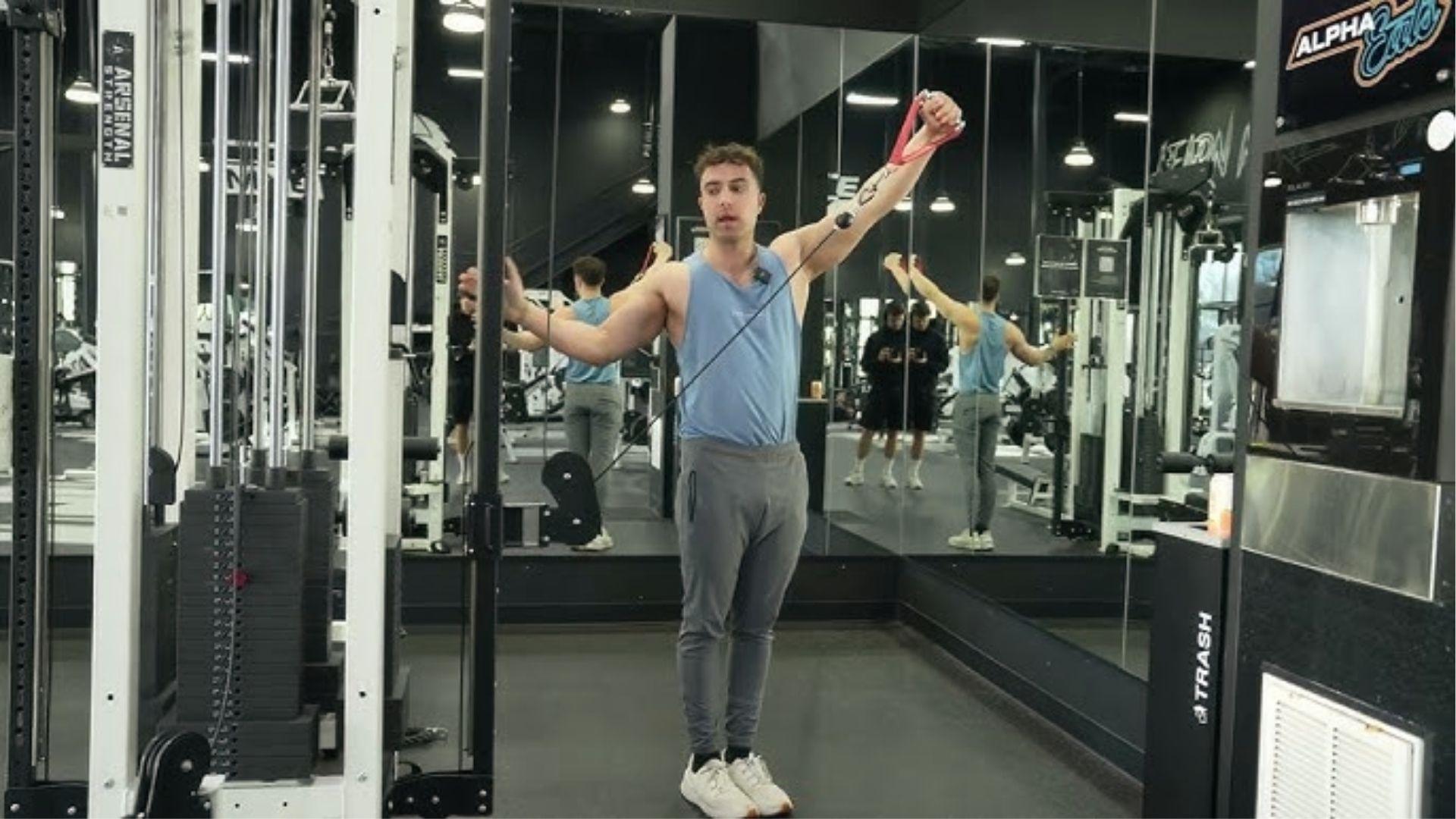 Man in blue tank does single-arm cable Y raise with resistance band grip in mirrored gym