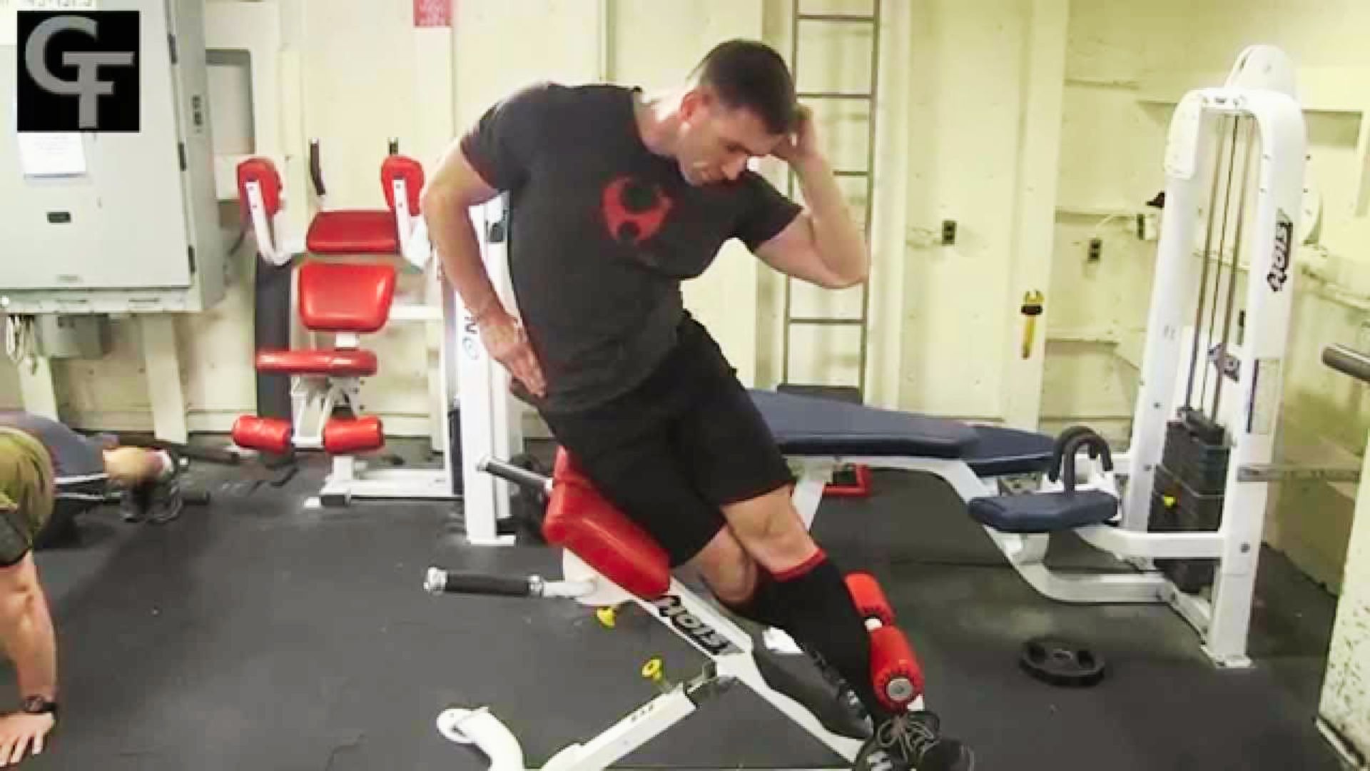 A man in a black t-shirt and shorts does a Roman chair side bend in a gym with red equipment.