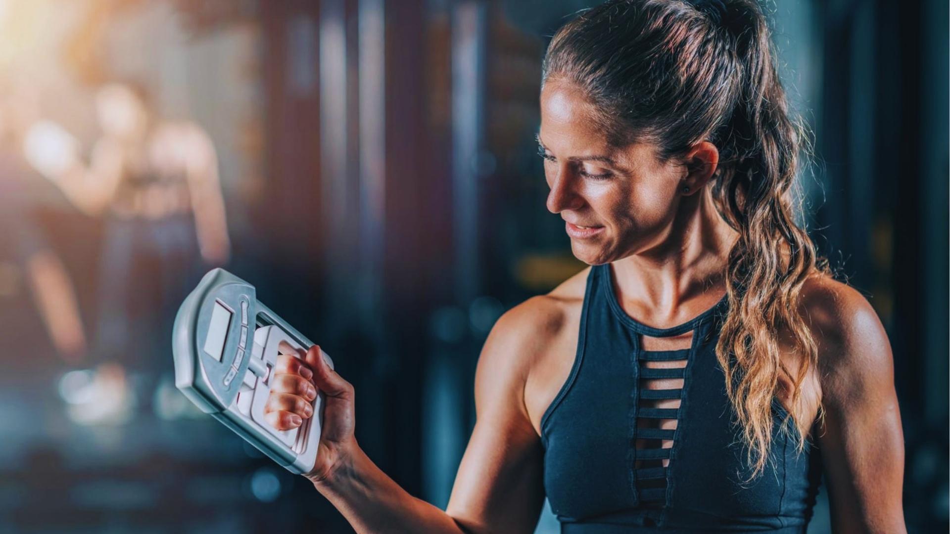 Athletic woman in black strappy tank top smiles while squeezing a digital hand dynamometer during strength test in gym.