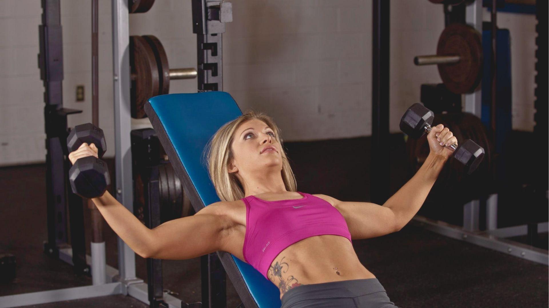 Woman in a pink sports bra lying on an incline bench, arms outstretched holding dumbbells to her sides.