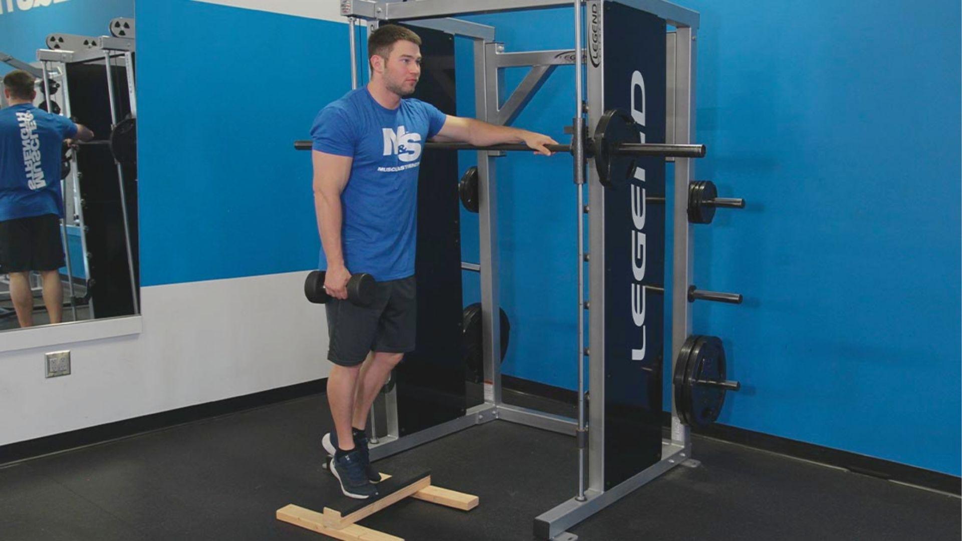 Man in blue Muscle & Strength shirt holds a dumbbell while standing on a wooden calf block next to a Smith machine.