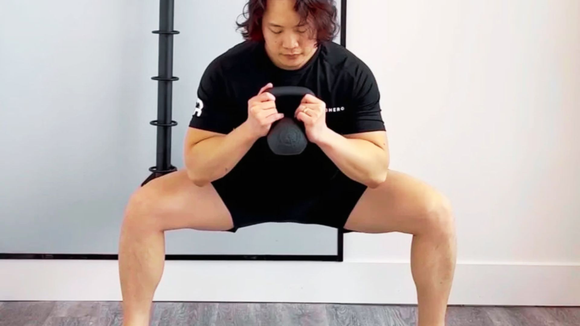 Man demonstrating a kettlebell sumo squat in a studio, gripping the weight with both hands at the bottom of the movement.