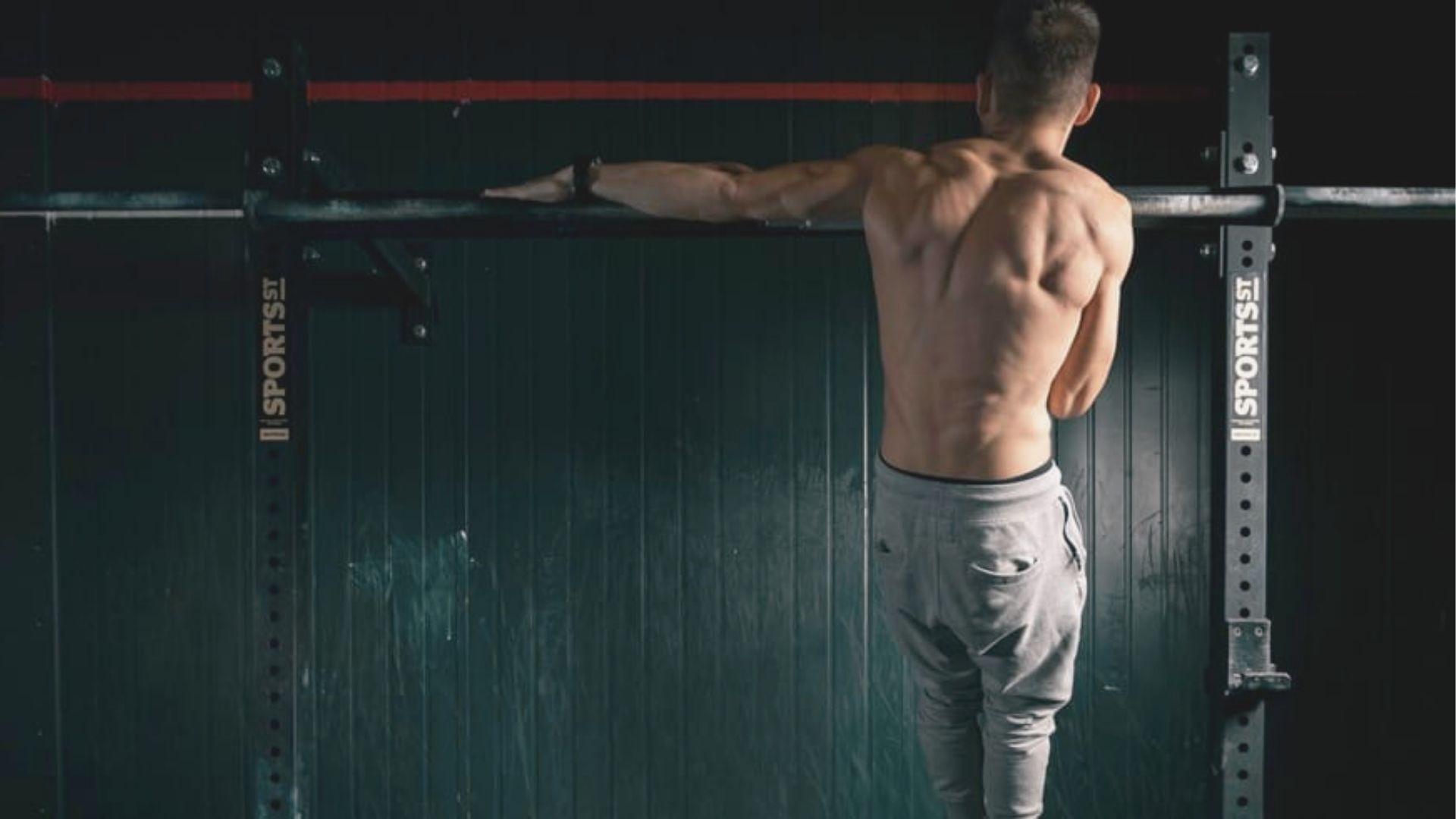 Shirtless muscular man in gray sweatpants doing a one-arm assisted pull-up on a squat rack bar.