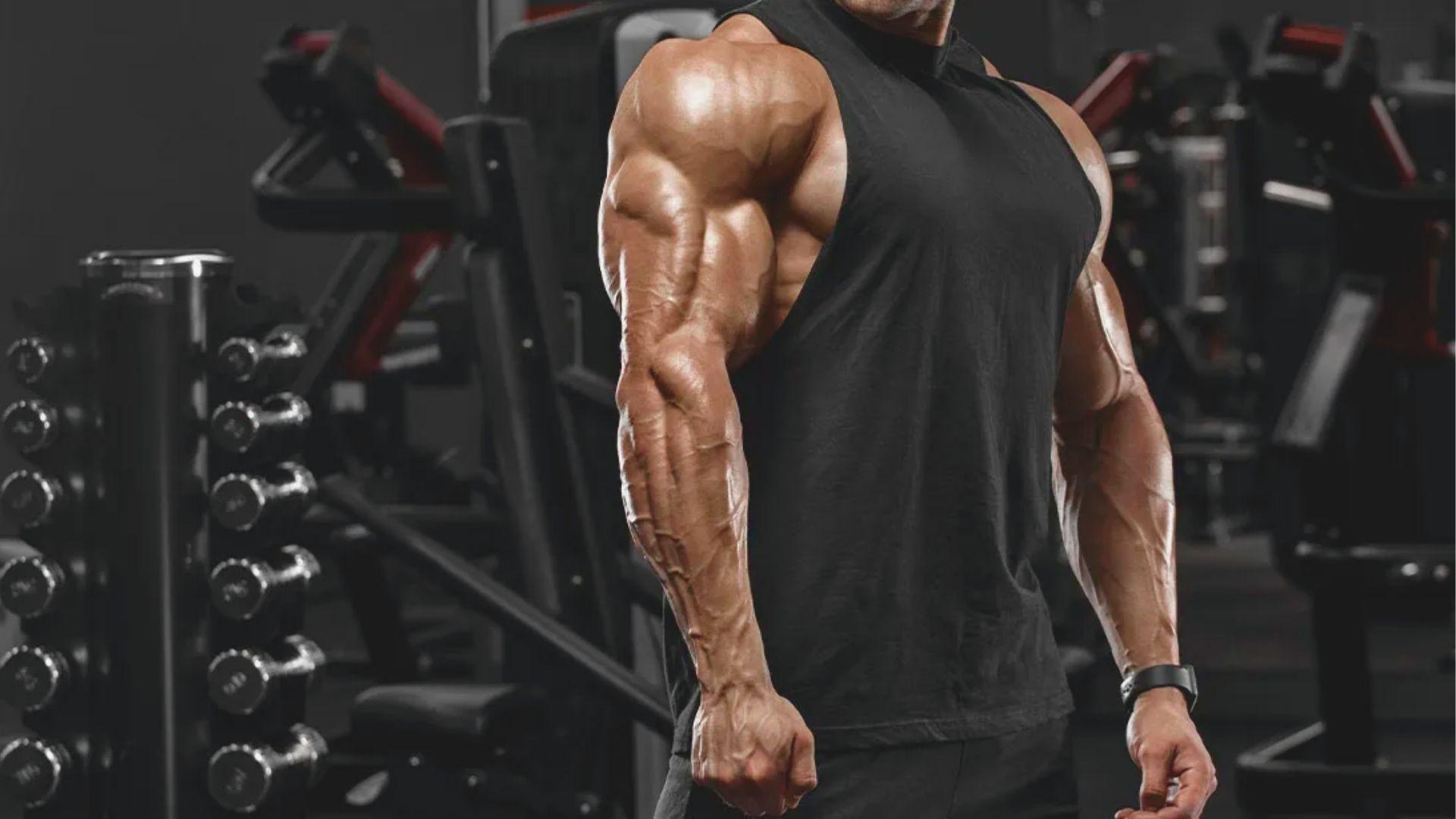 Muscular man in black tank top flexing triceps and shoulder muscles in a gym.