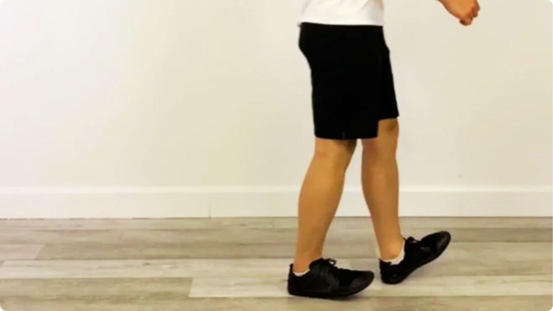 Person in black shorts and sneakers walking on heels with toes lifted off the floor indoors.