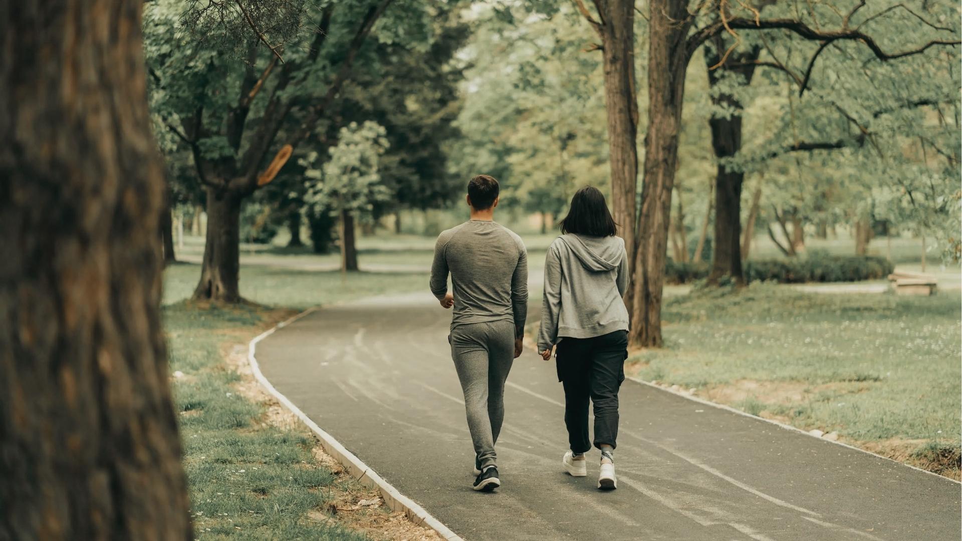 Man and woman in gray activewear walk away on curved park paths lined with tall trees for daily steps.
