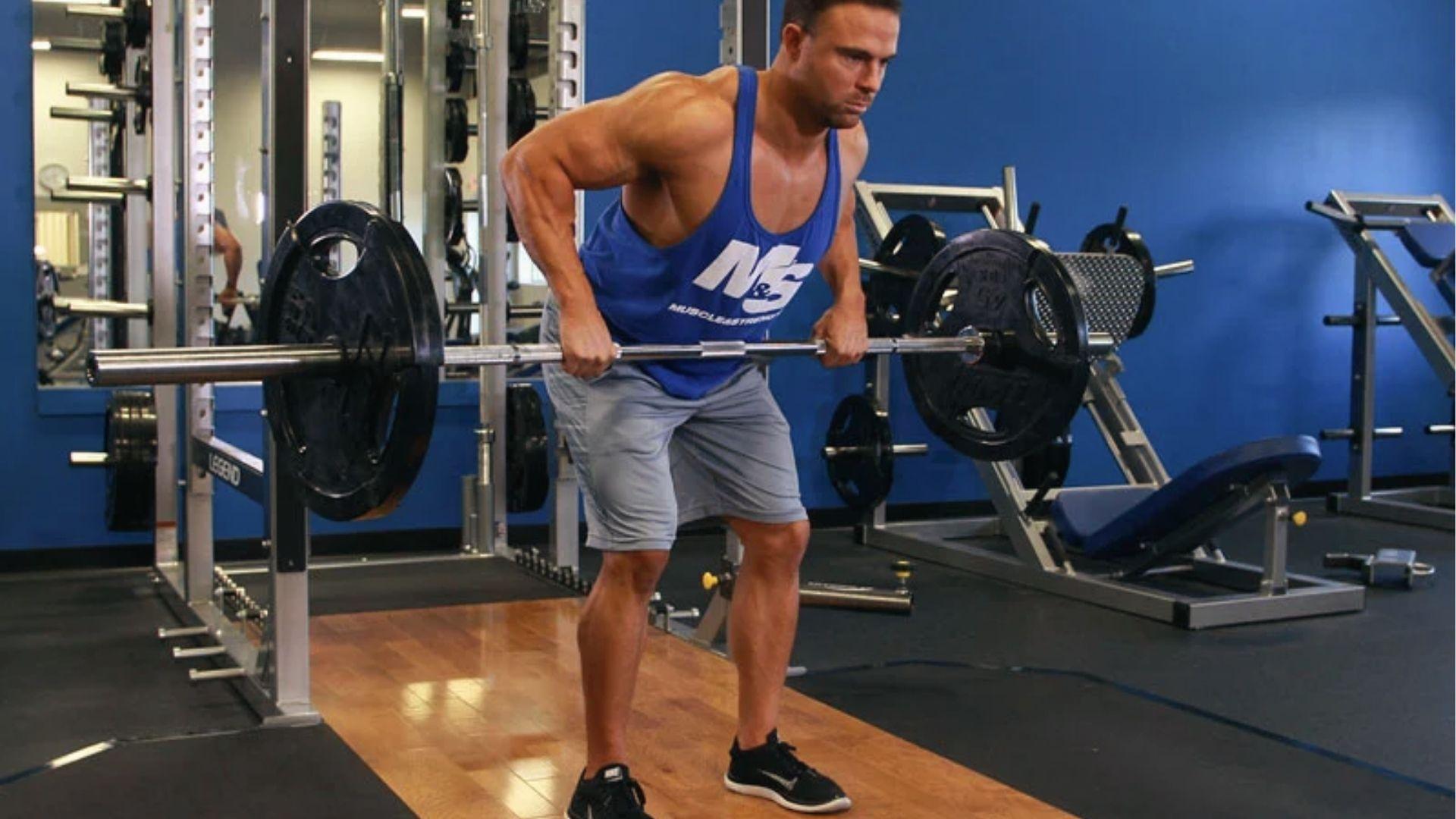 Man in blue tank top demonstrates bent over barbell row form with wide grip in fitness gym