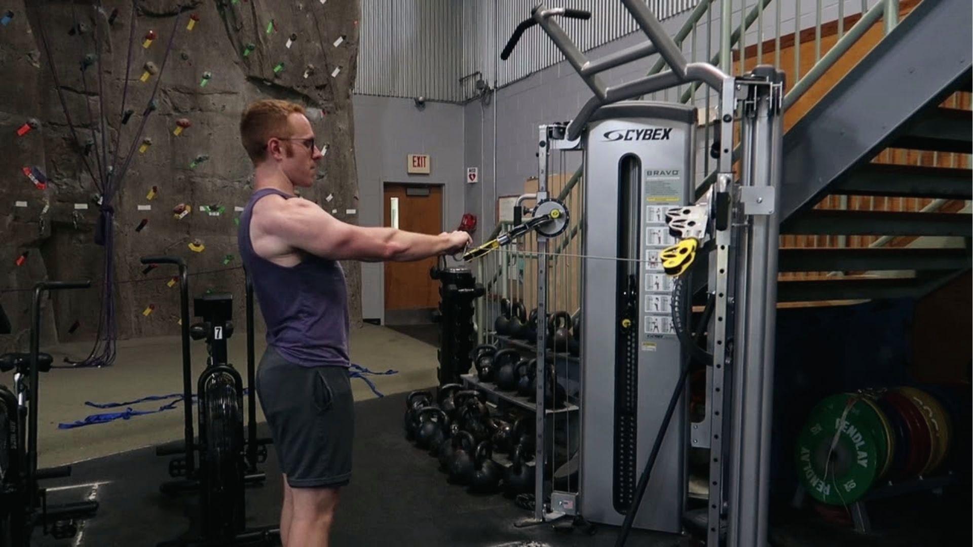 Man in purple tank demonstrates cable Y raise form on Cybex machine near climbing wall.
