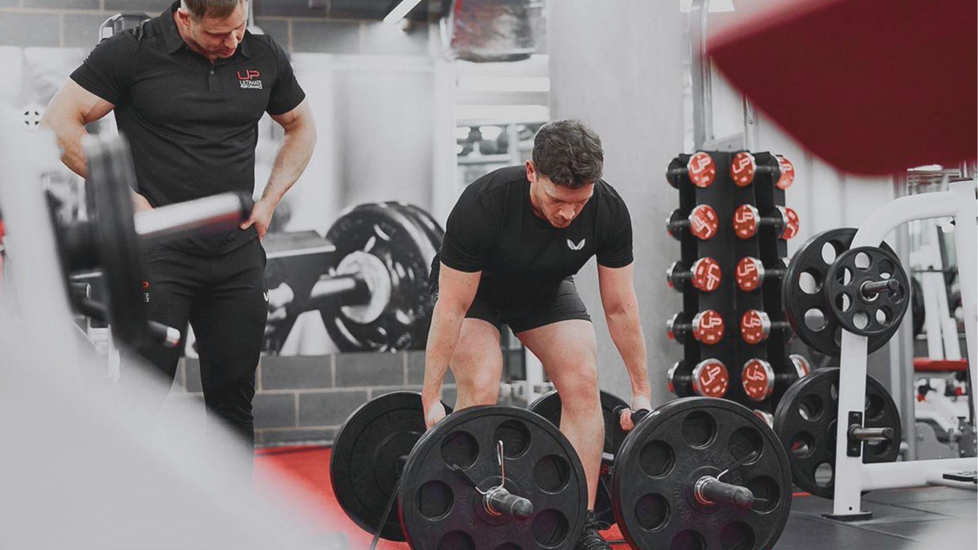 Man gripping a loaded trap bar in a gym, preparing to lift, while a trainer watches closely.