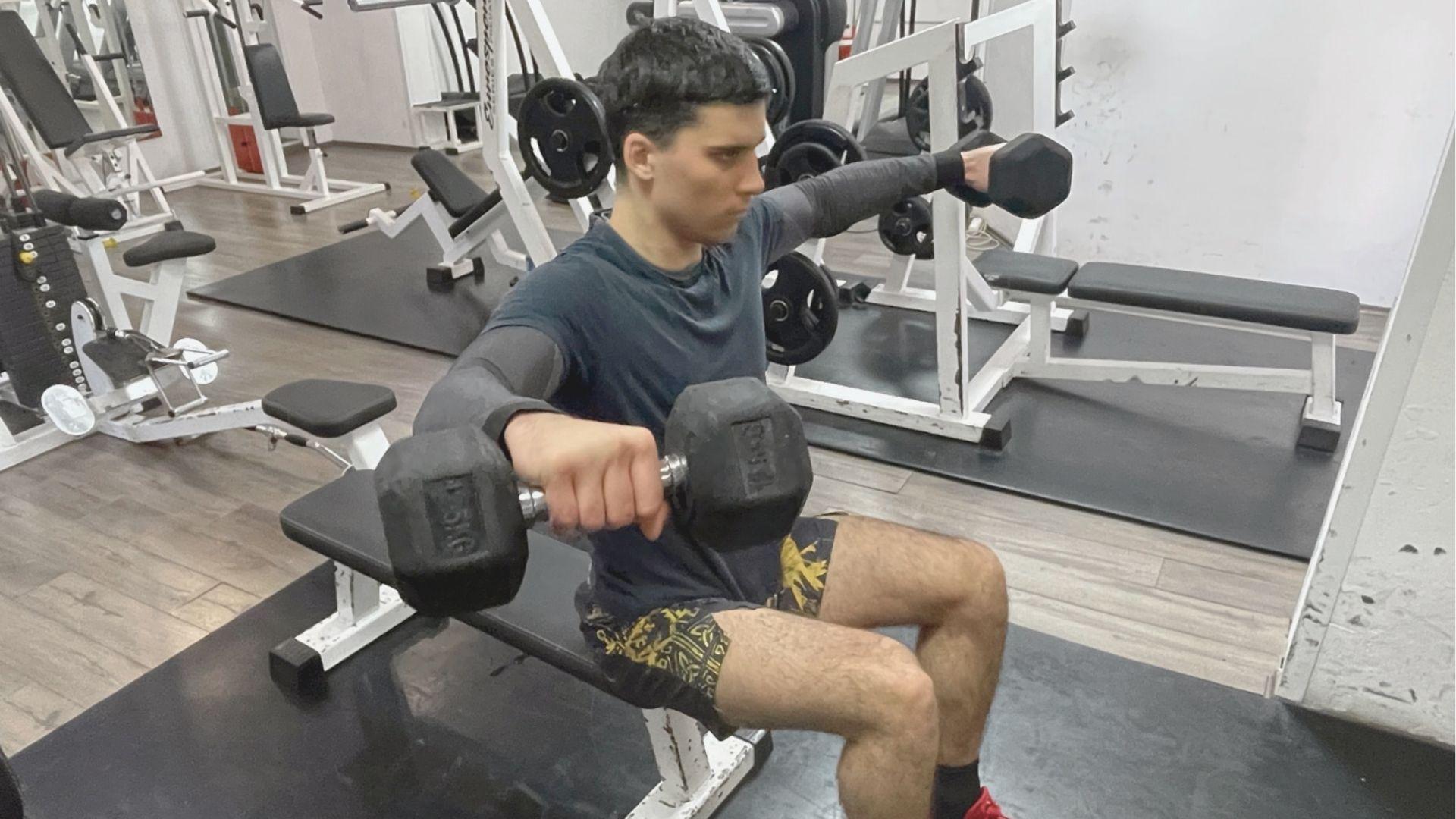 A man seated on a gym bench executes dumbbell lateral raises with arms parallel to the floor.
