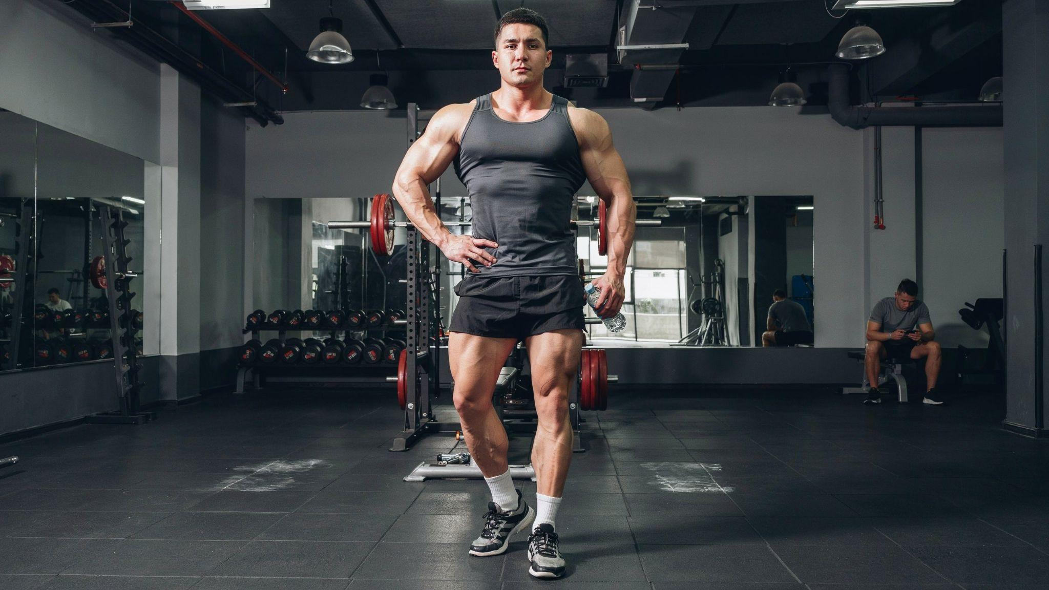 Fit man poses by barbell in gym, chest and arms built but quads and calves look disproportionately slim.