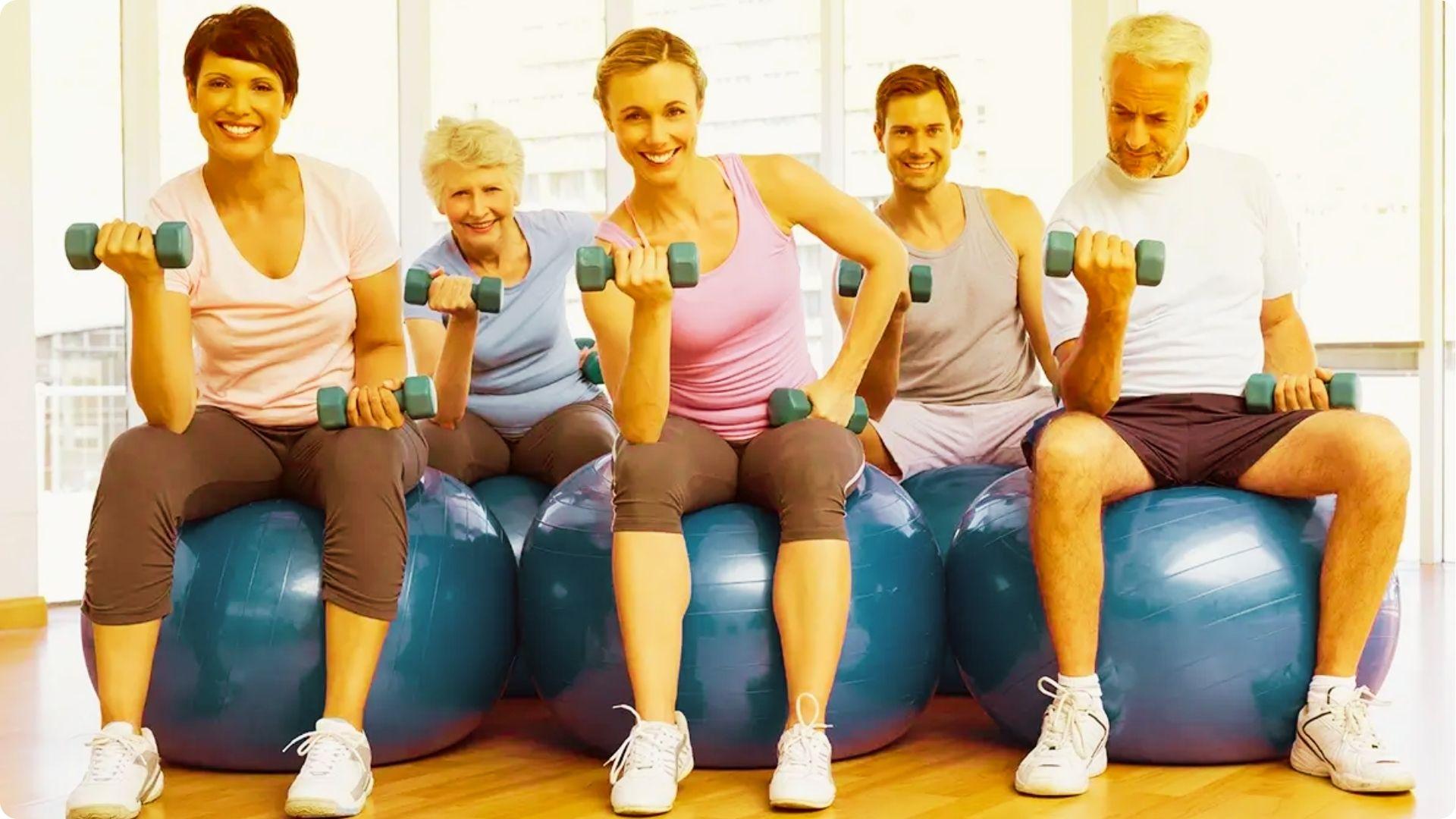 Five adults of different ages, from young to senior, sit on exercise balls doing dumbbell curls in a bright gym.