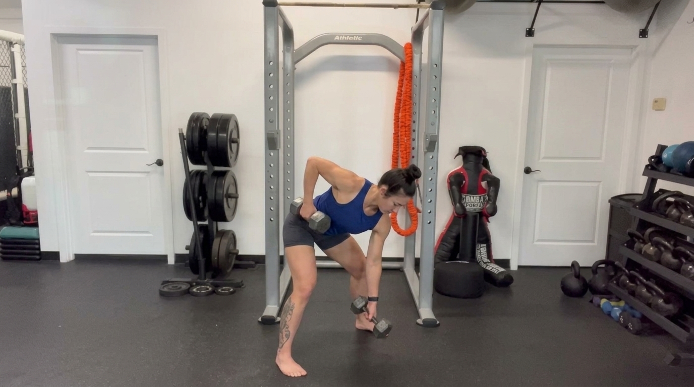 A woman in a blue top does a bent-over dumbbell row with staggered stance in a home gym.