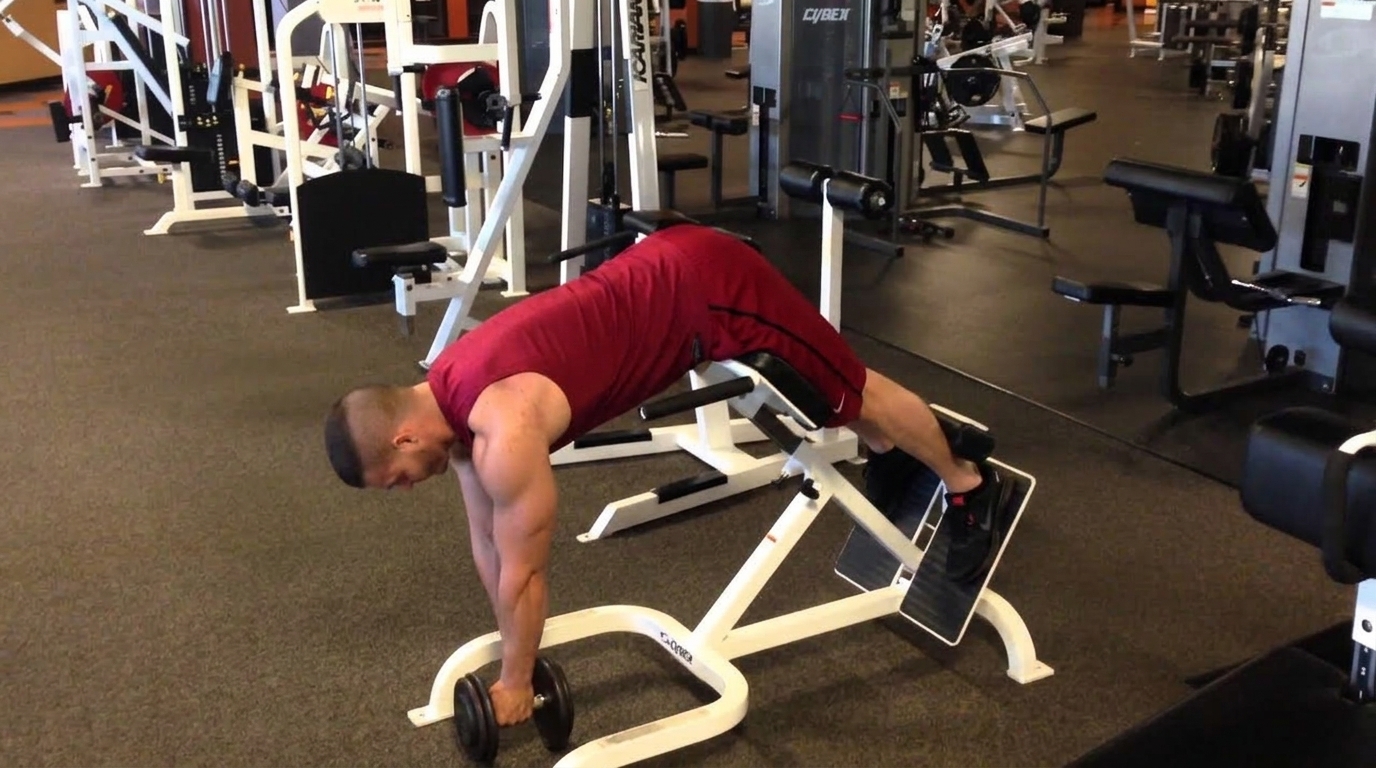 Man in red tank top performs a weighted back extension on a 45-degree hyperextension bench, gripping a dumbbell.