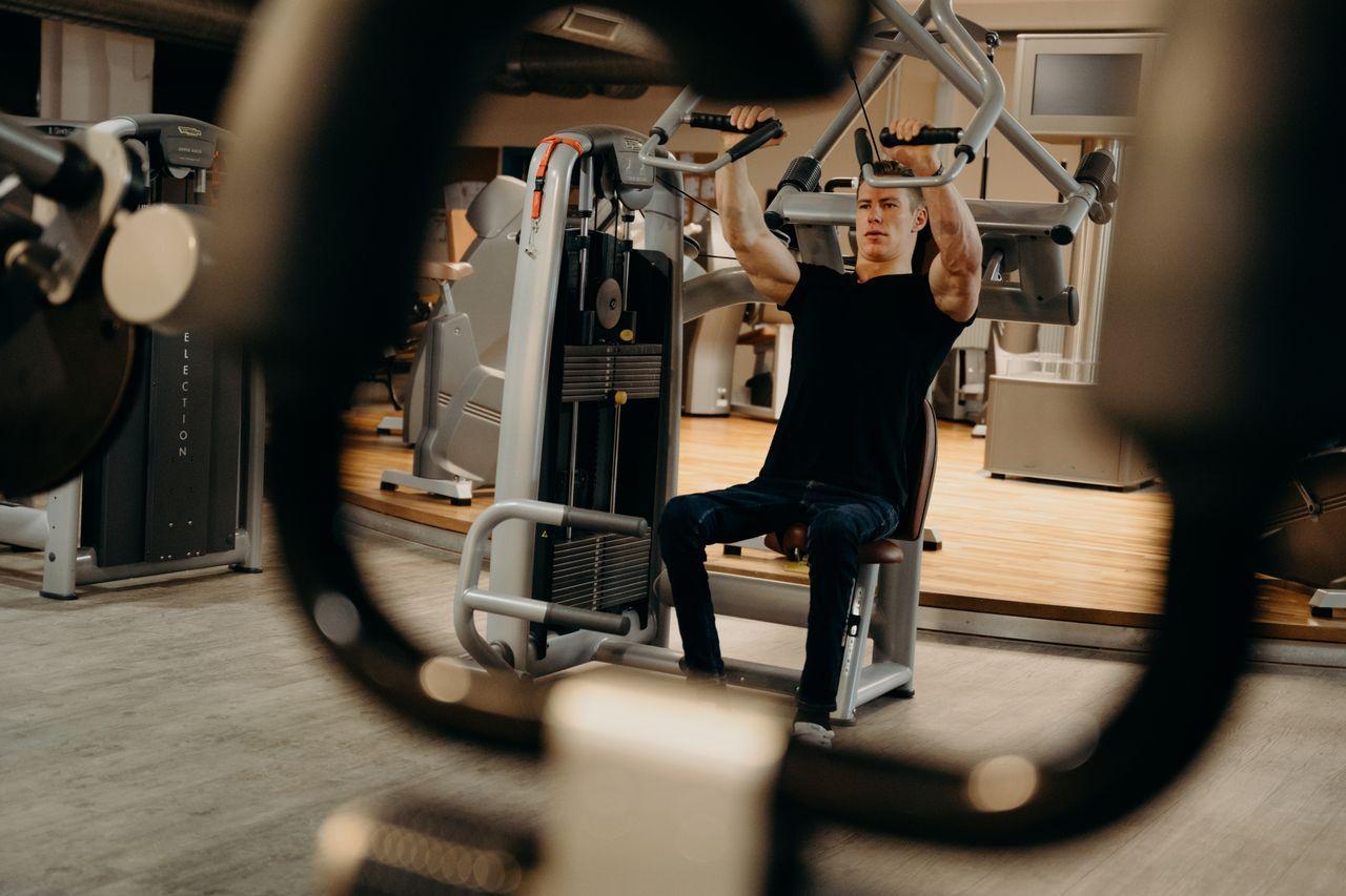 A man seated at a chest press machine in a modern gym, viewed through the equipment frame in the foreground.