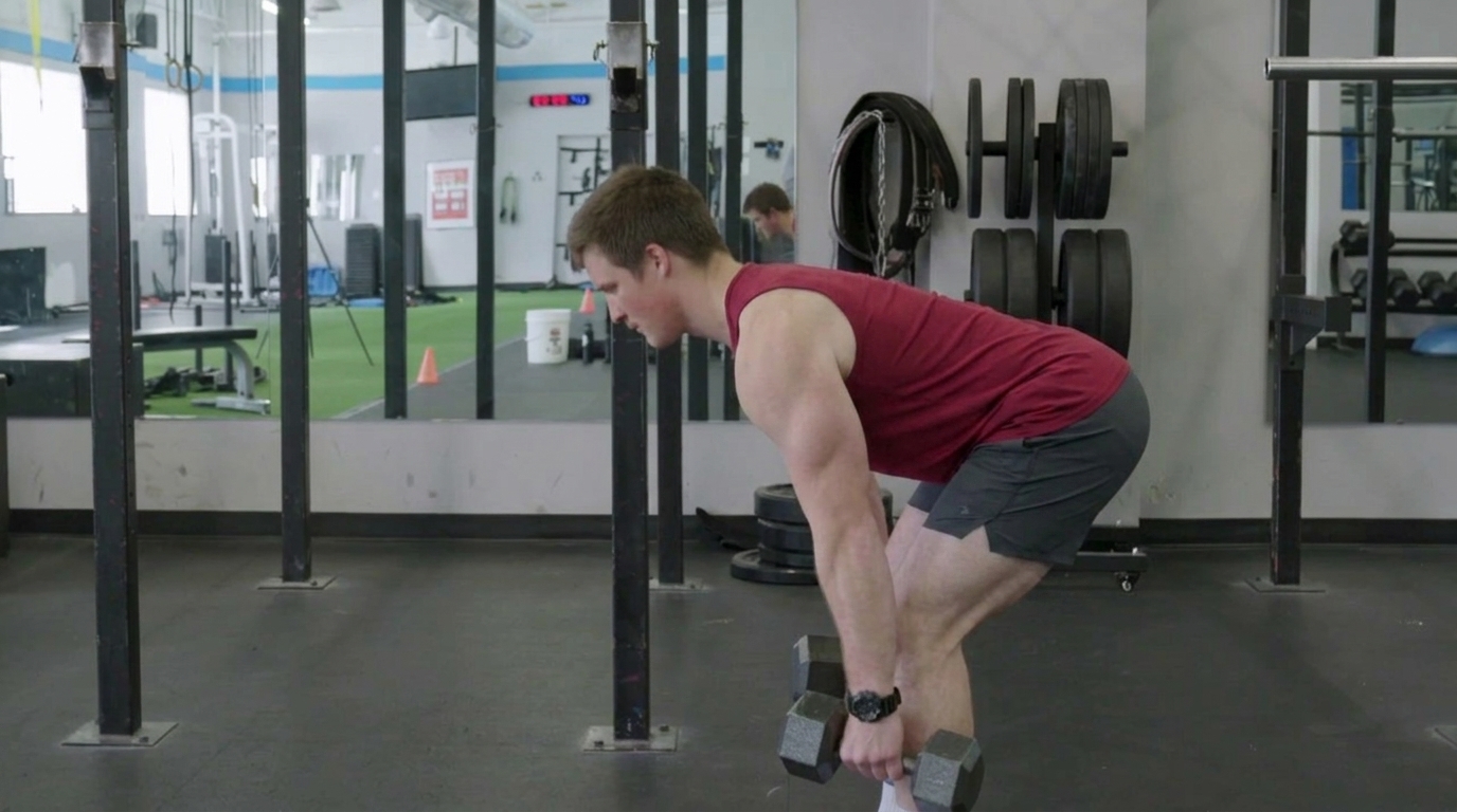 Man in red sleeveless shirt executes a dumbbell Romanian deadlift in a gym with weight racks behind him.