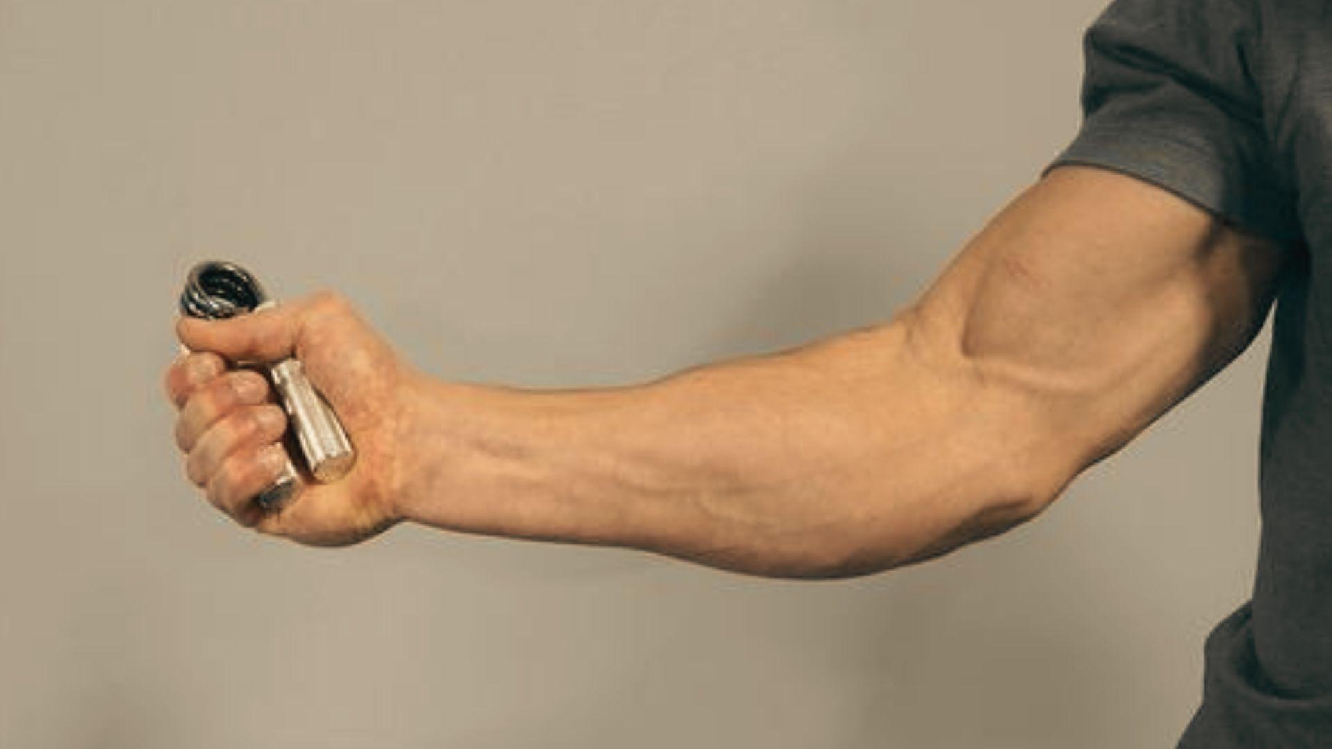 Muscular man squeezing hand gripper device to strengthen forearms, veins visible, wearing black shirt, athletic field background