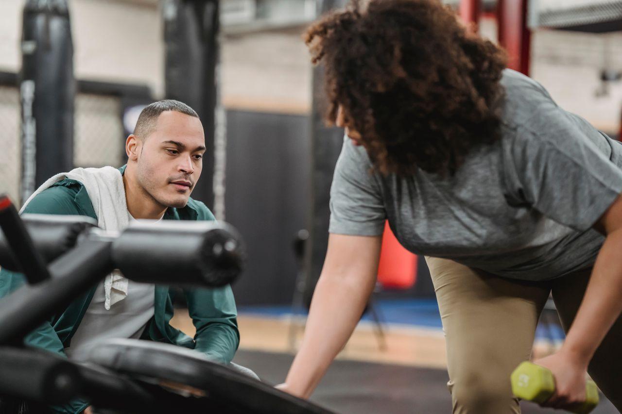 A focused trainer watches a client perform a dumbbell exercise during a one-on-one session in a gym.