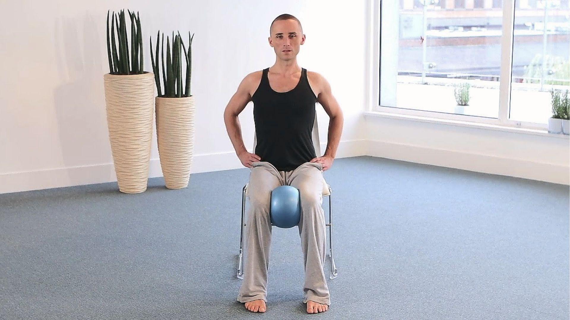 A man in black tank top sits on a chair squeezing a blue ball between knees for an inner thigh workout.