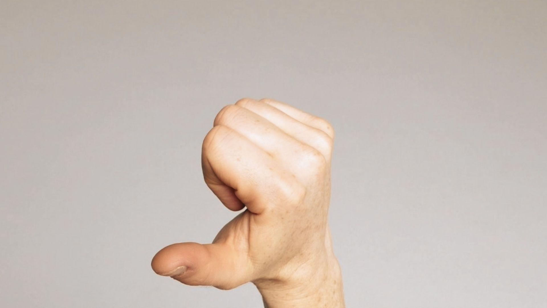 Close-up of right hand making closed fist with thumb extended downward, signaling thumbs down gesture, plain background