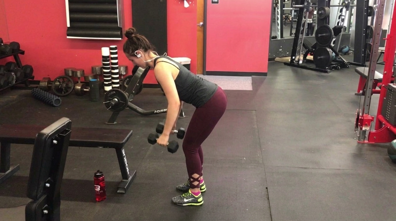A woman in maroon leggings performs bent-over dumbbell rows in a gym with red walls.