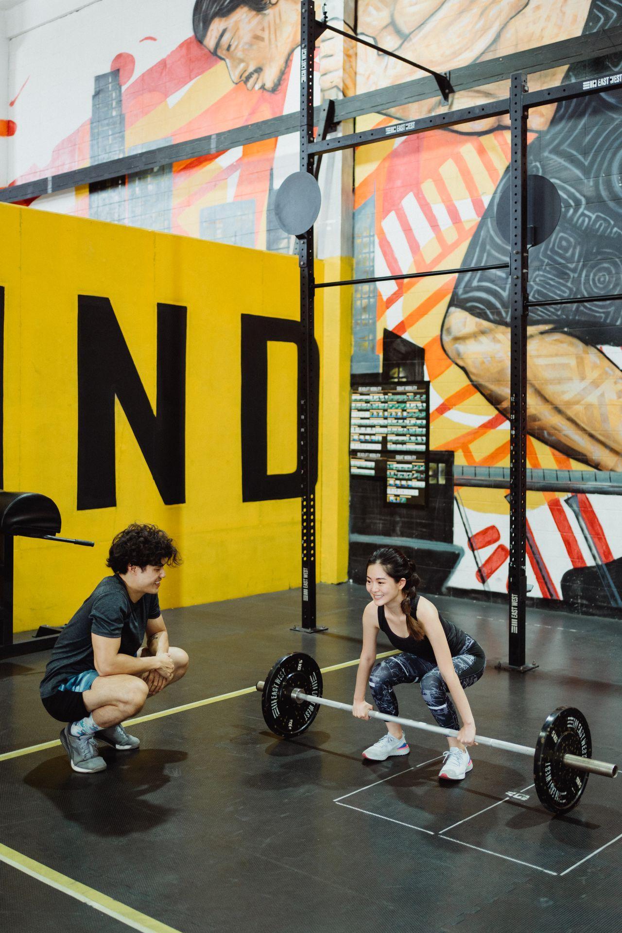 A trainer crouches beside a smiling woman as she sets up for a barbell deadlift in a colorful gym.