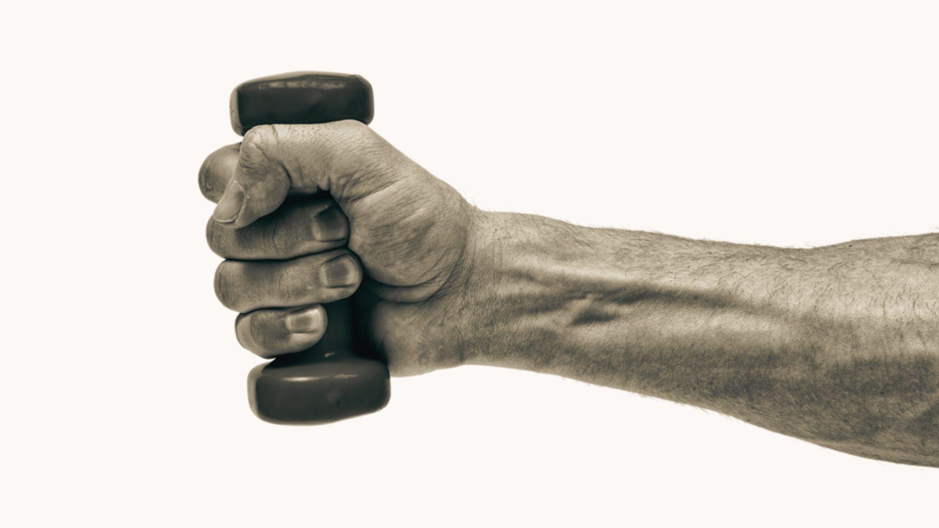 Sepia-toned close-up of muscular forearm and hand gripping small black dumbbell tightly, veins prominent, white background, strength exercise
