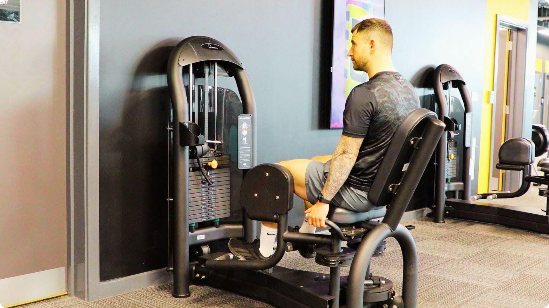 Man seated on a hip abduction machine in a gym, pushing legs outward against resistance pads.