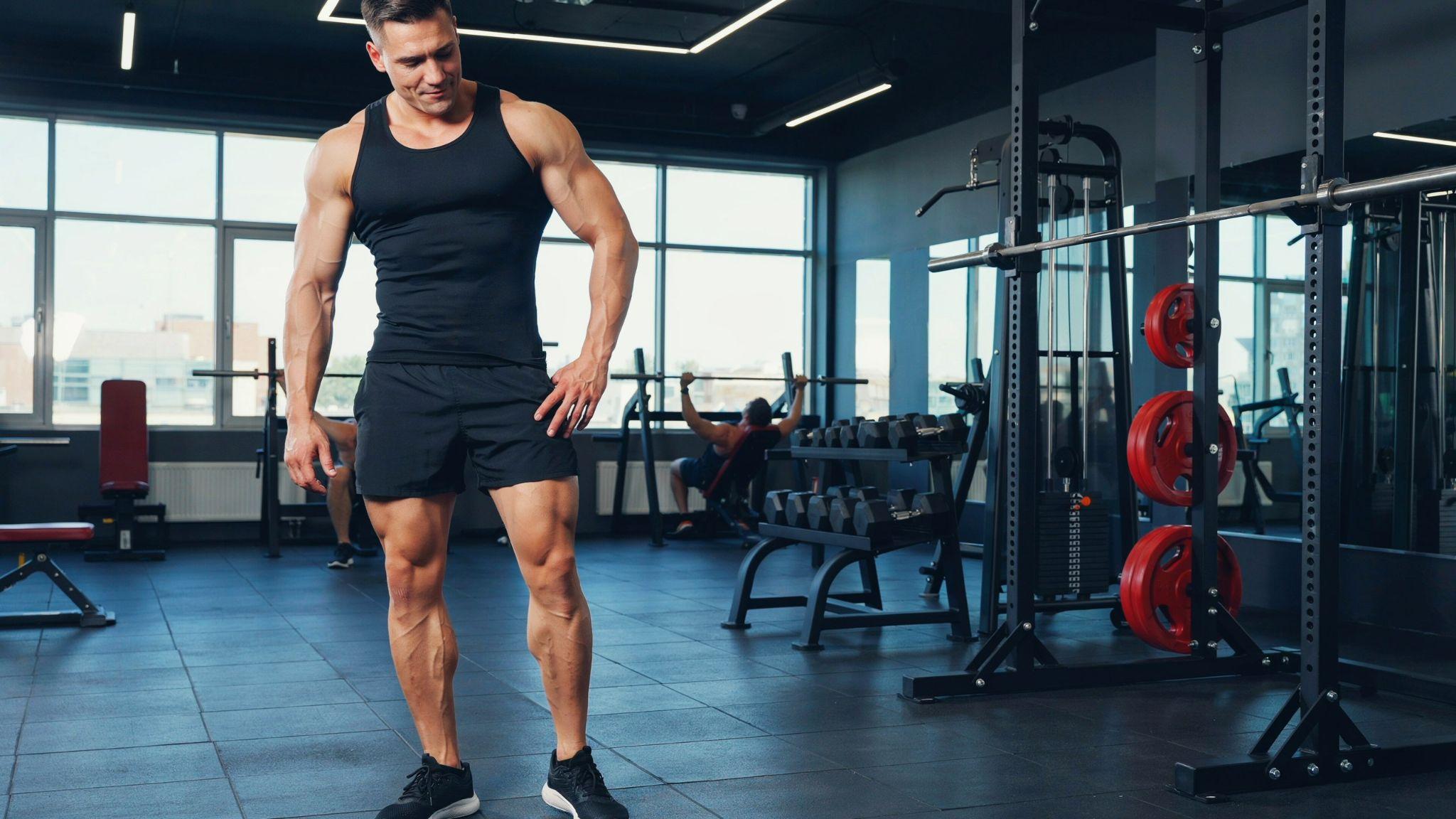 Muscular man in tank top stands in gym with bulky upper body and noticeably underdeveloped legs.