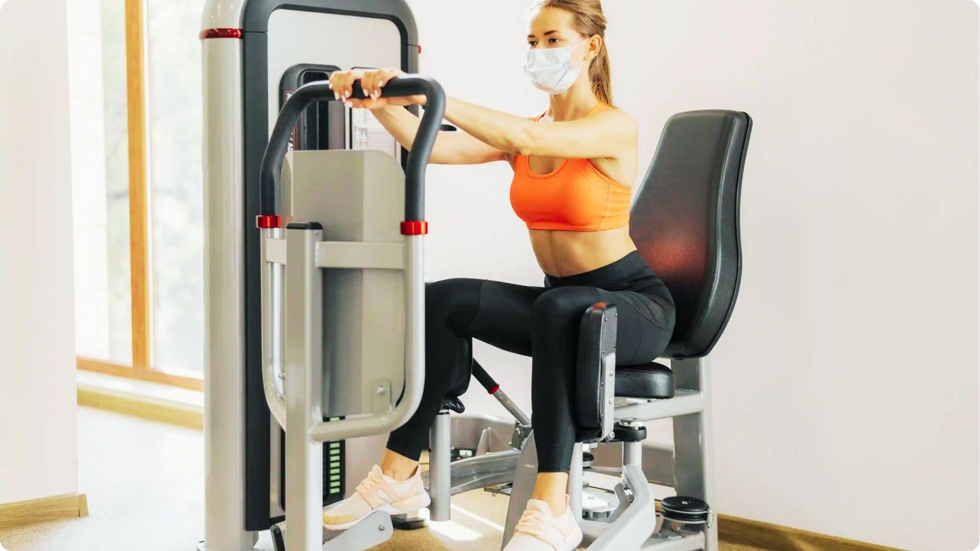 Woman wearing a face mask uses a seated hip abduction machine, holding handles while pressing thighs outward.