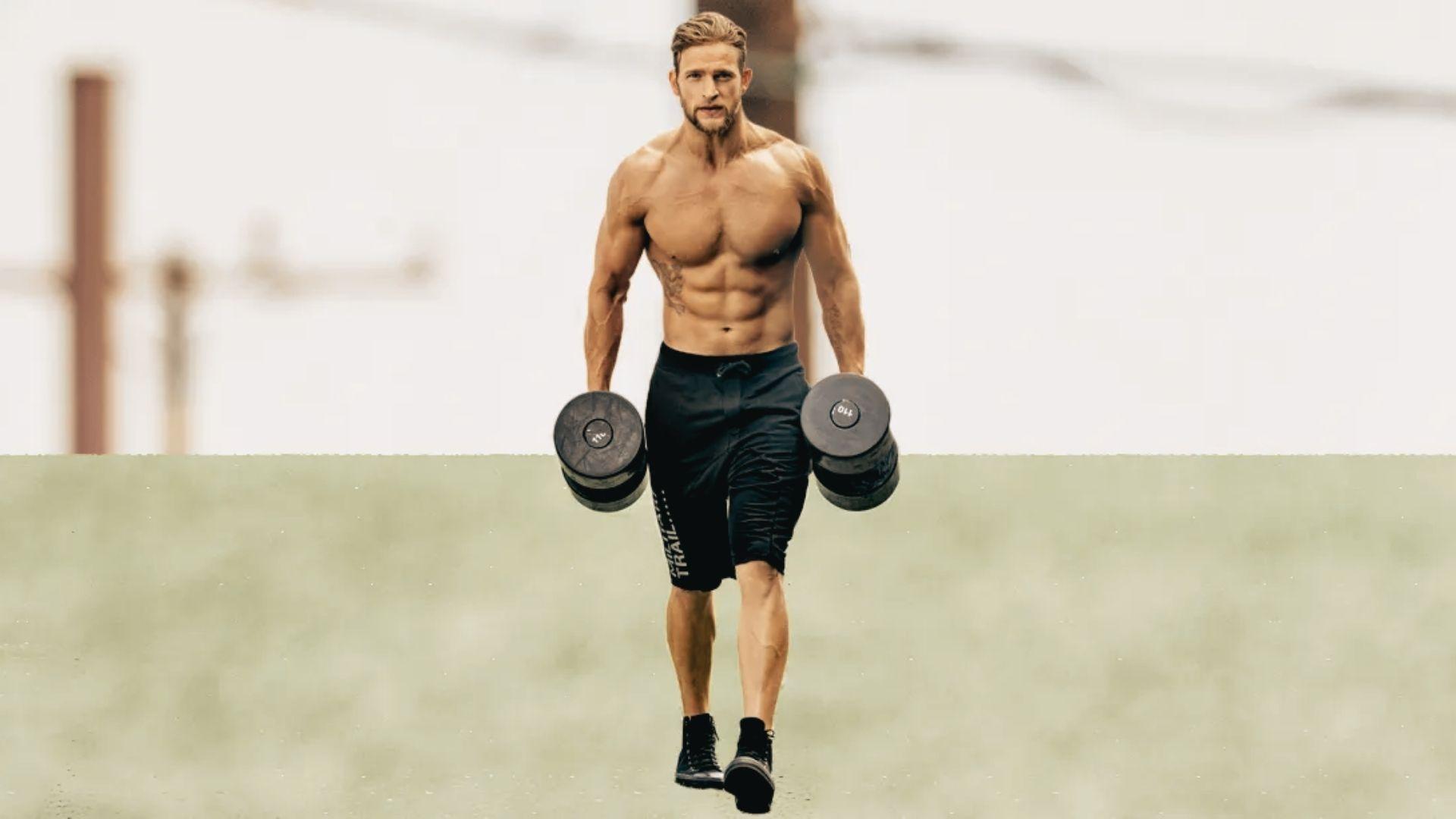 Shirtless muscular man walking outdoors performing farmer’s carry with heavy dumbbells in each hand, black shorts, blurred background
